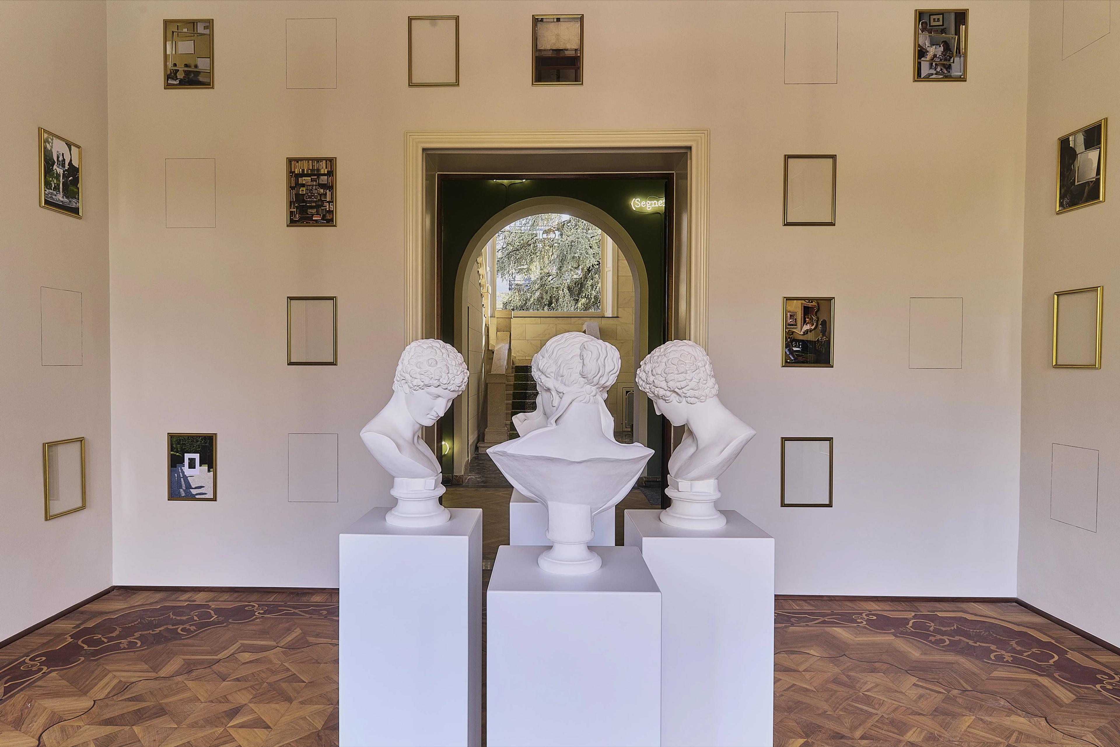 A hallway with three marble busts on pedestals, empty gilt frames on the walls, leading to a lit archway.