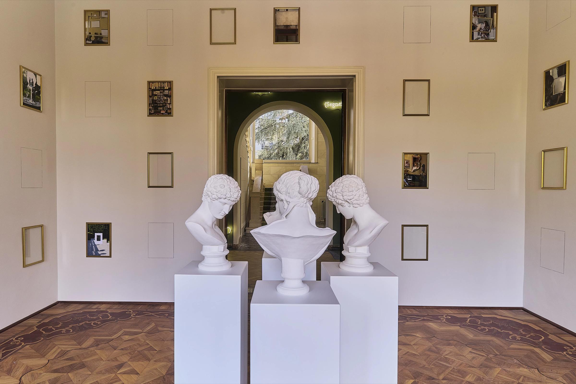 A hallway with three marble busts on pedestals, empty gilt frames on the walls, leading to a lit archway.