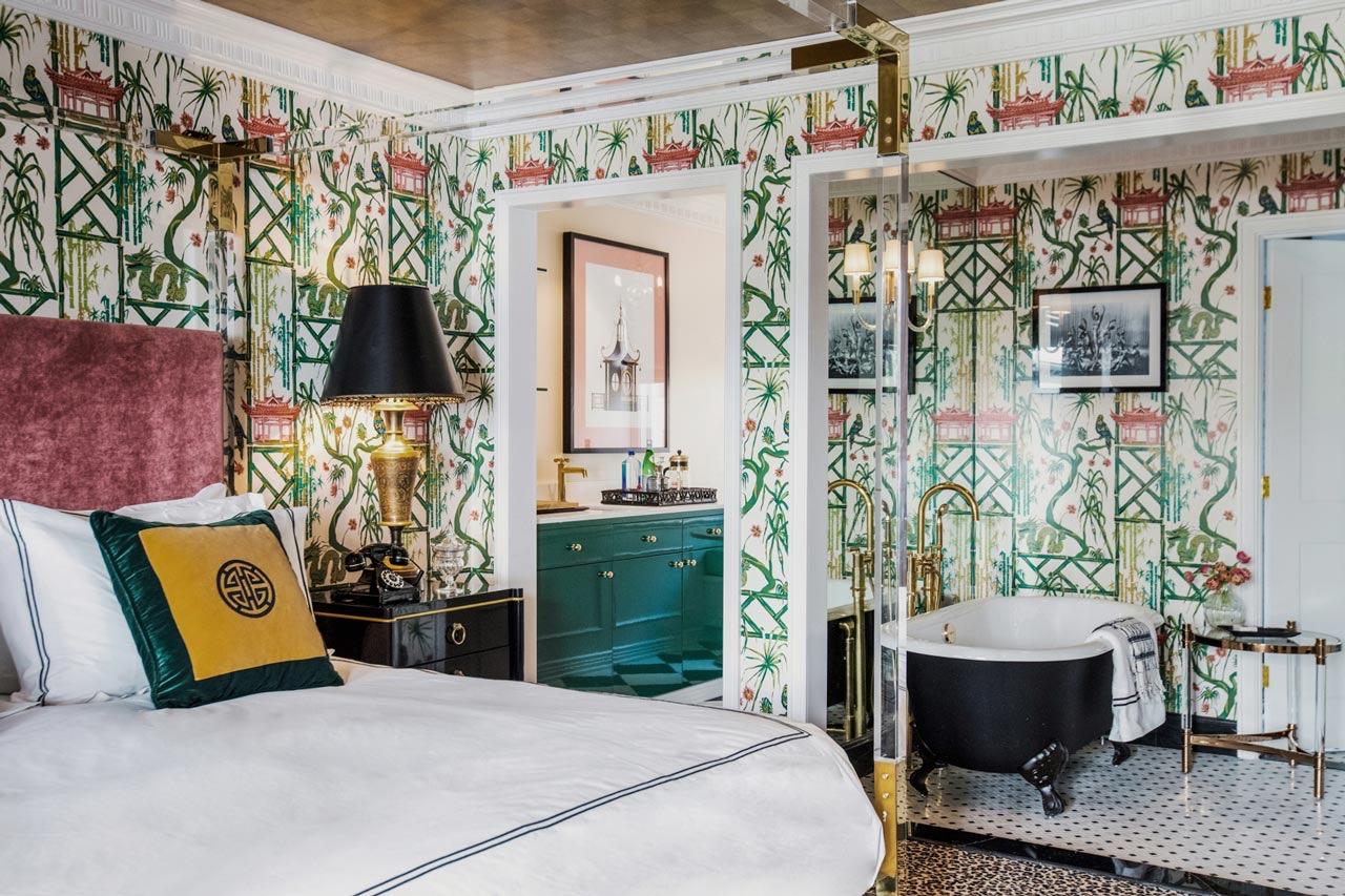 A bedroom and bathroom divided by a glass partition, with green bamboo wallpaper, a maroon headboard, and a black clawfoot tub on black and white tiled flooring.