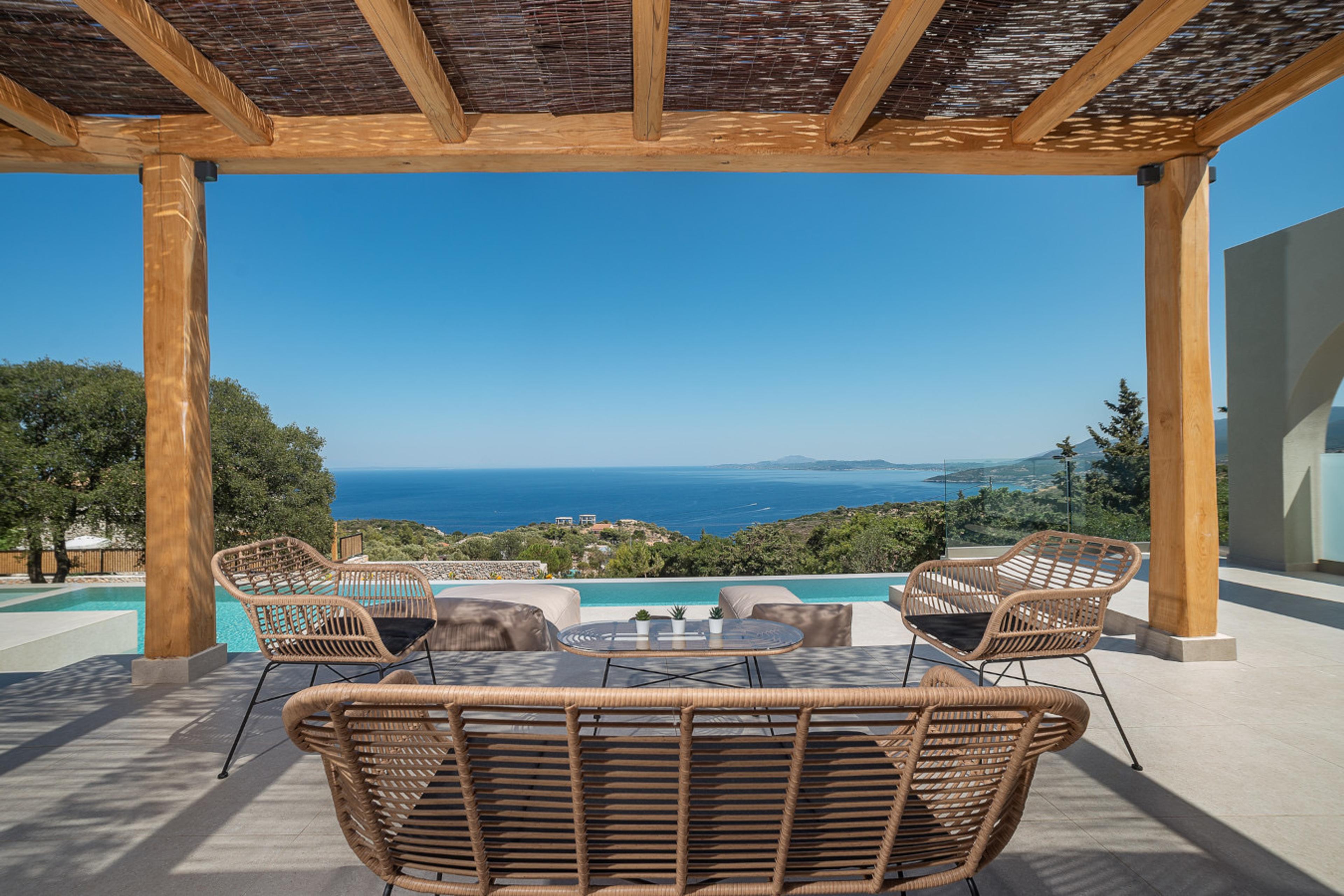 Woven rattan chairs and a sofa under a pergola, facing an open sea view with hills along the coastline.