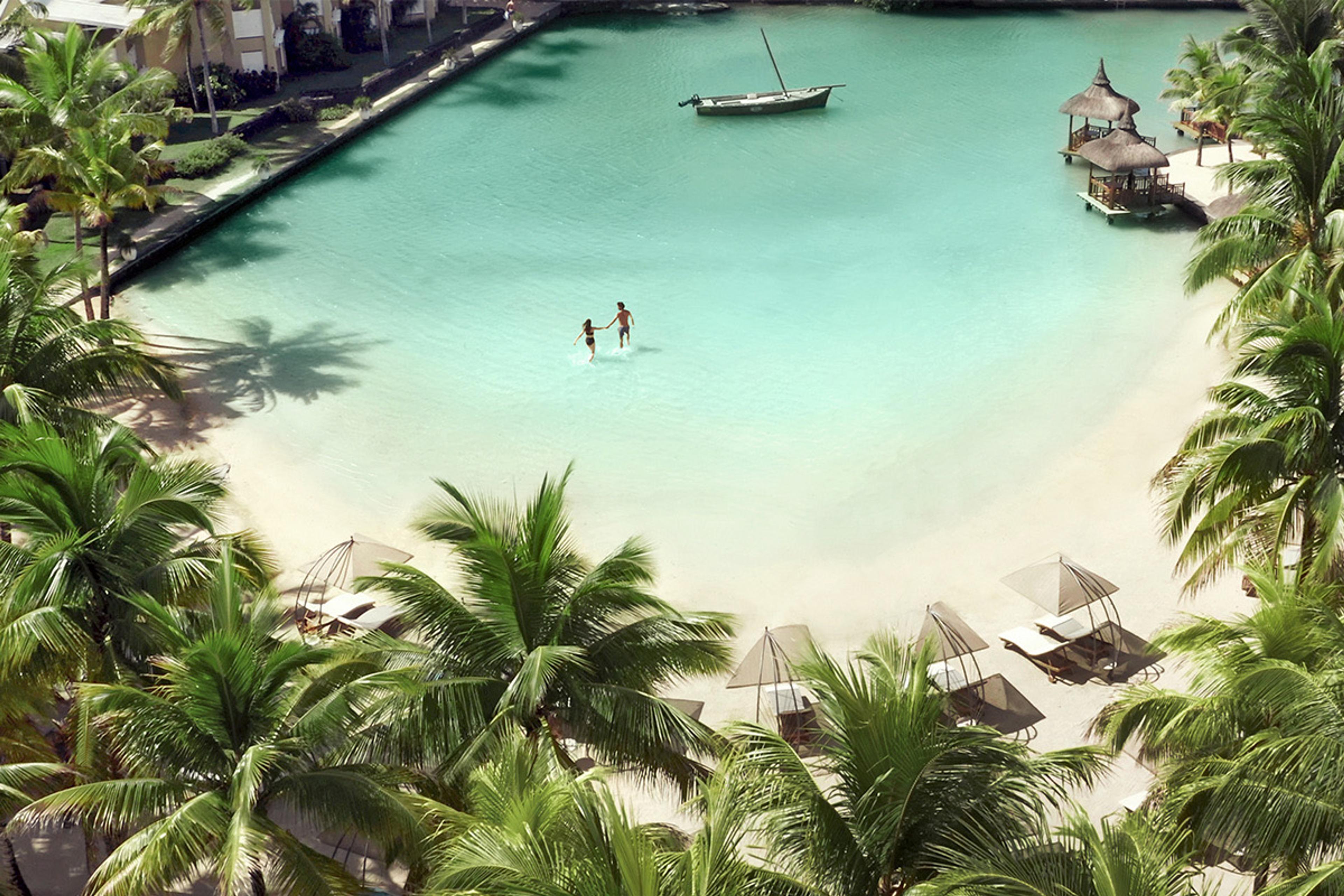 An aerial view of a turquoise lagoon with a wooden boat and thatched sunshades along a white sand shore, framed by palm trees, with two people wading in the shallow water.