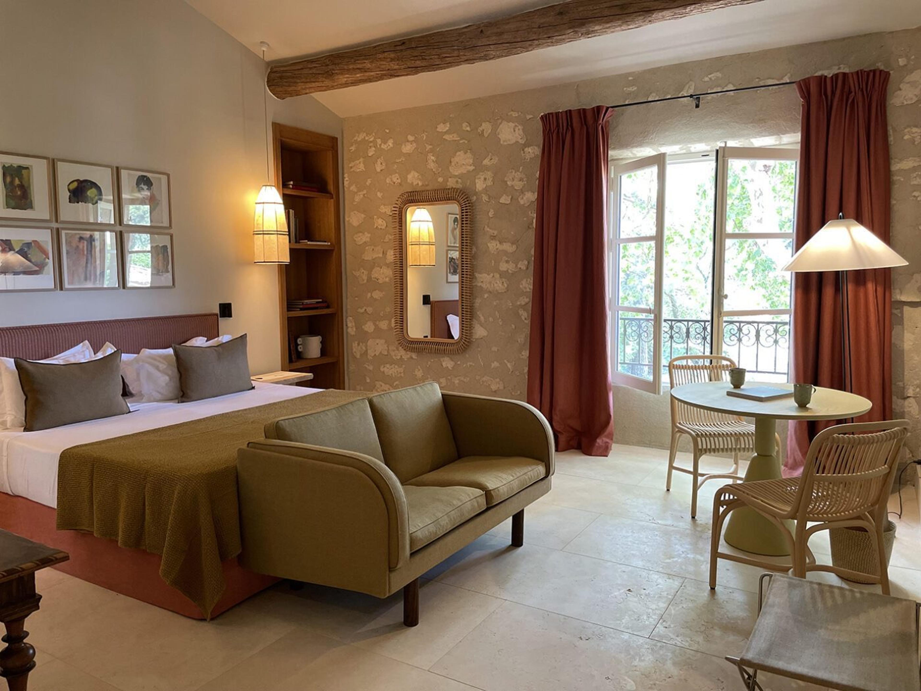 A hotel bedroom with a bed and an olive sofa facing a round breakfast table, with a balcony behind red curtains.