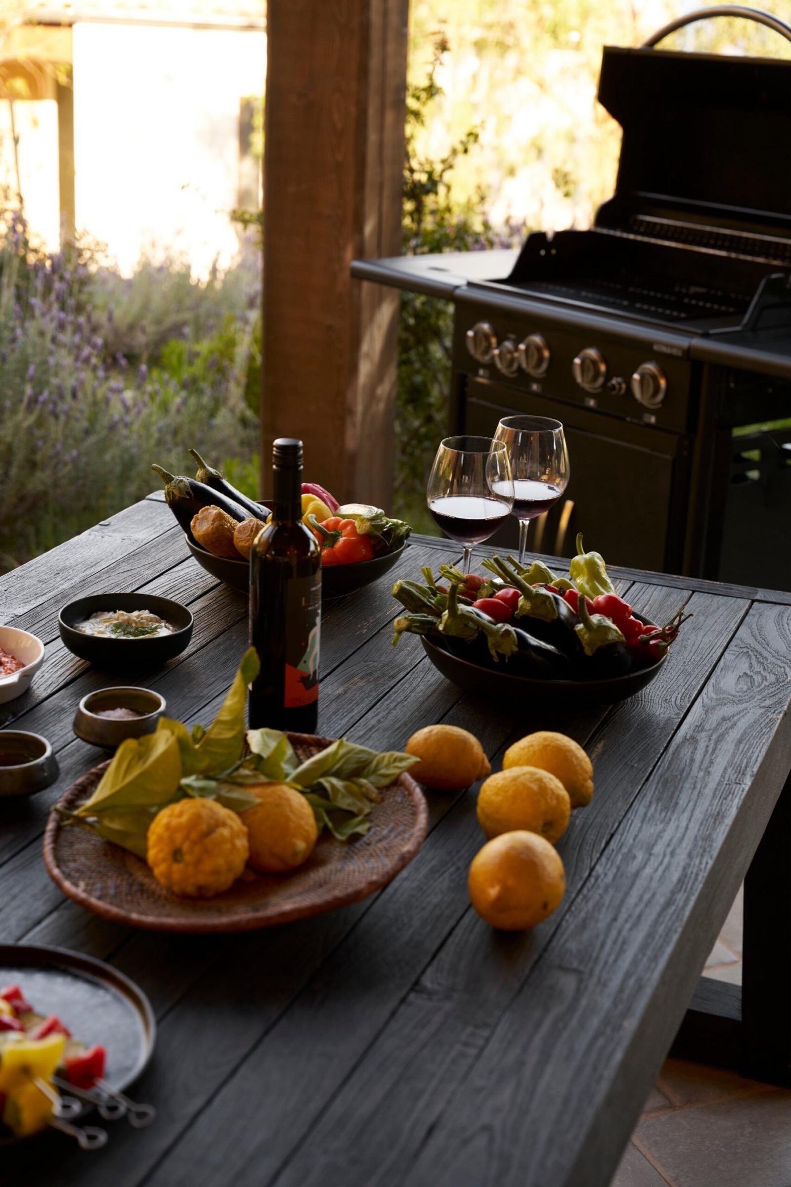 a wooden outdoor table laid with bowls of vegetables, a plate of lemons, two glasses of red wine and an open wine bottle, a barbecue behind it