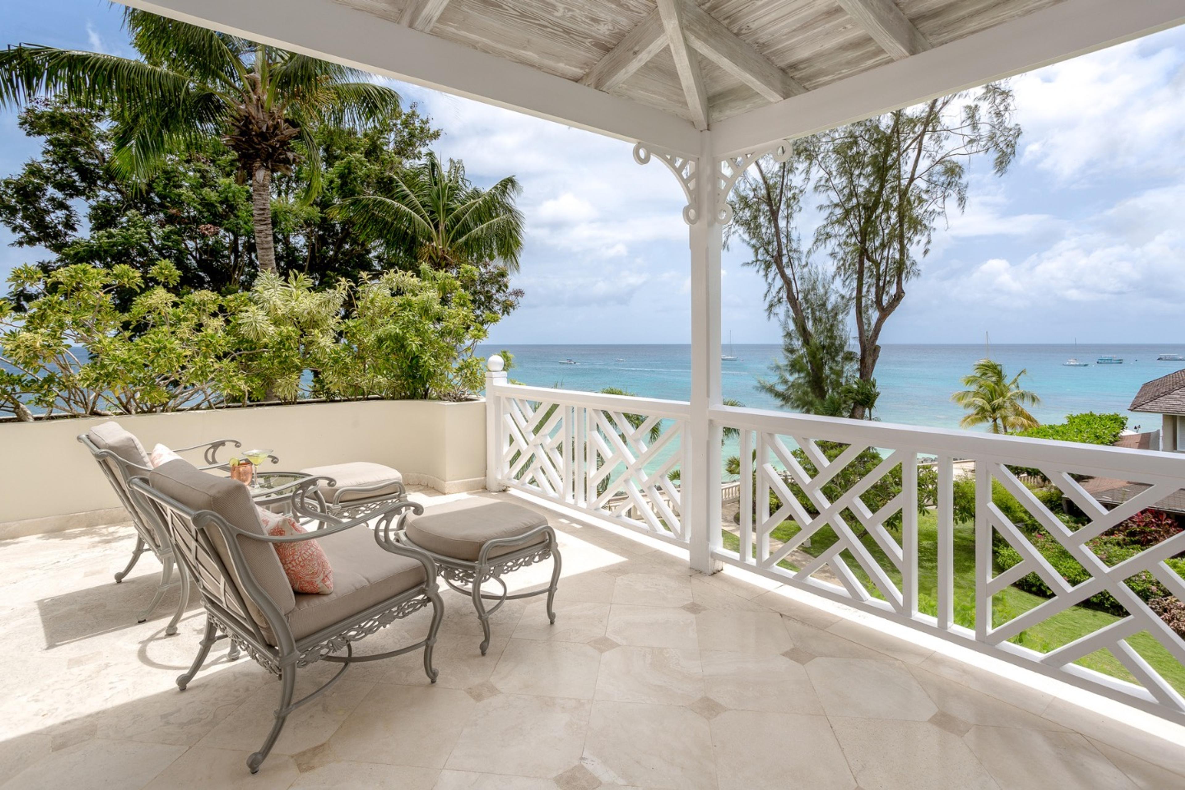 A covered balcony with cushioned wicker loungers and a footstool, framed by palm trees and a white balustrade, looking out over the sea.
