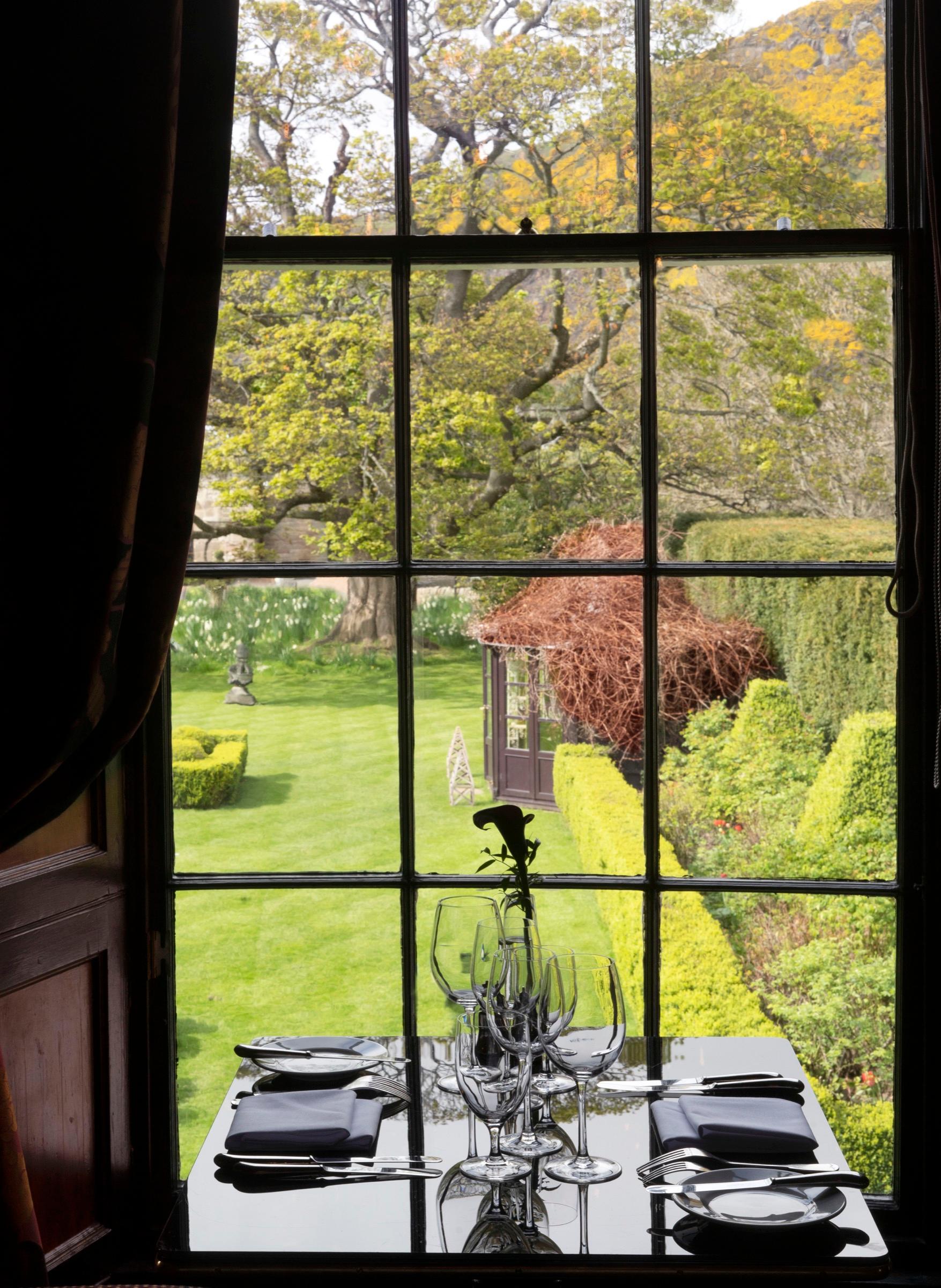 a table set for two beside a tall window looking out over a lawn, clipped hedges, a summerhouse and a hill beyond