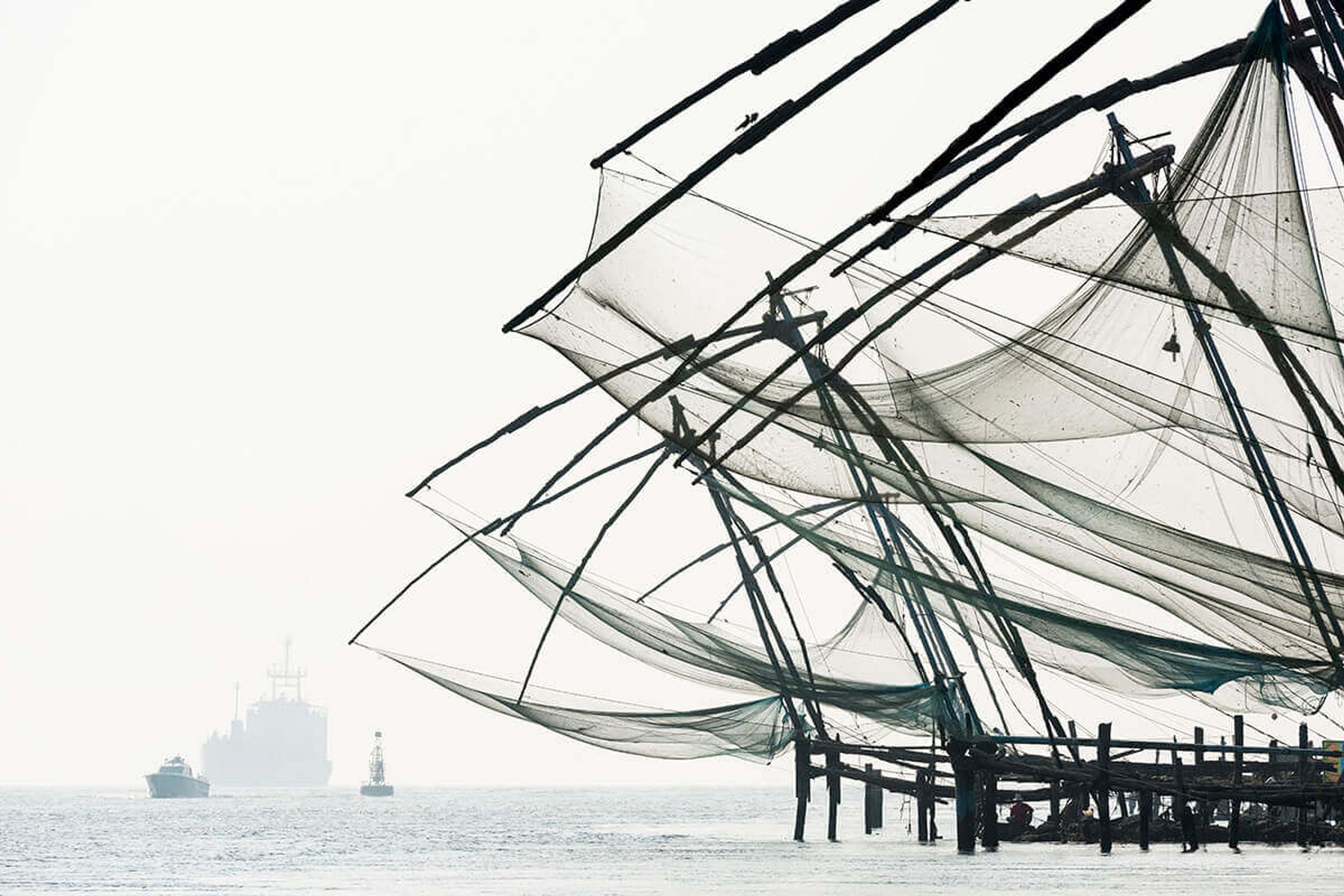 Wooden cantilevered fishing nets on a misty waterfront, with a cargo ship and a small boat passing in the distance.