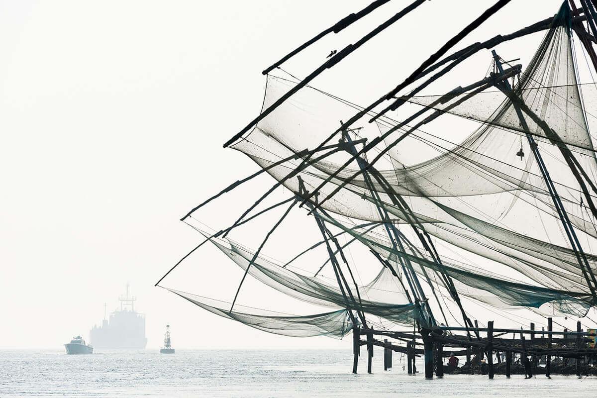 Wooden cantilevered fishing nets on a misty waterfront, with a cargo ship and a small boat passing in the distance.