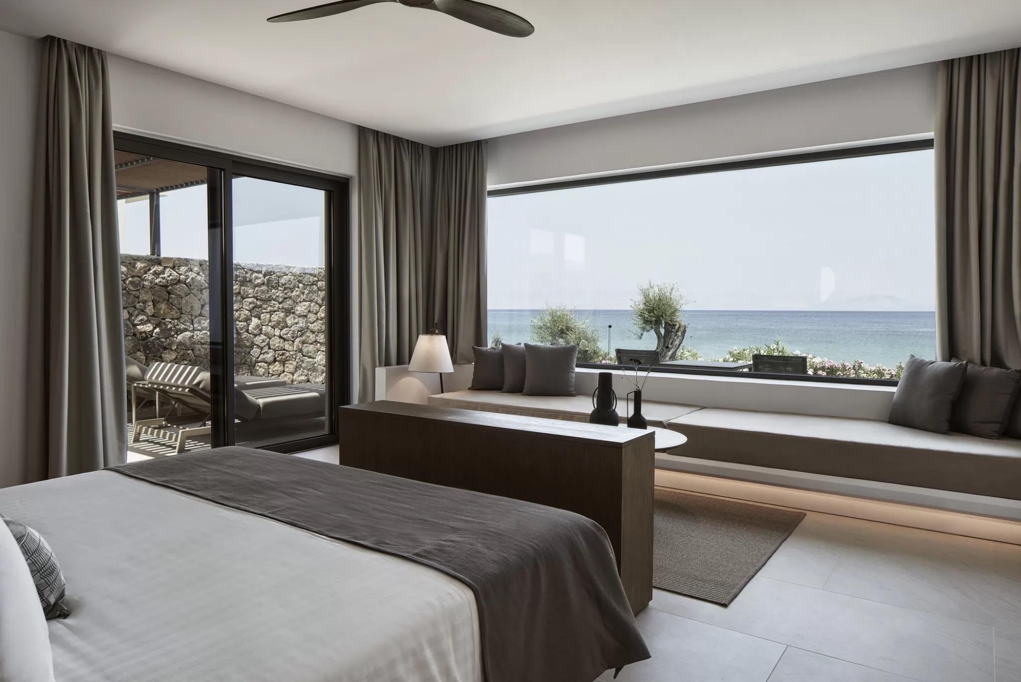 A hotel bedroom with a bench seat under a wide window facing the sea, and a bed with grey and white linen.