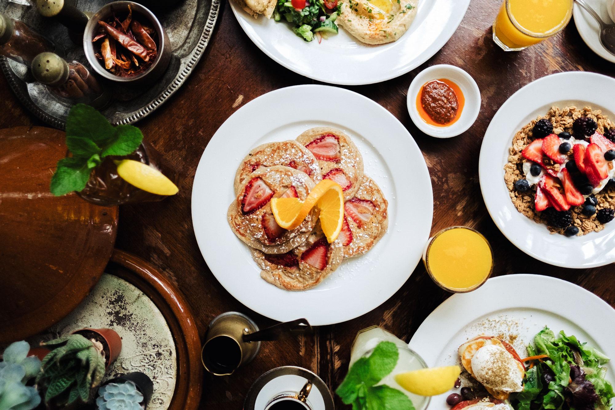 A brunch table seen from above, with pancakes, eggs, salads and orange juice