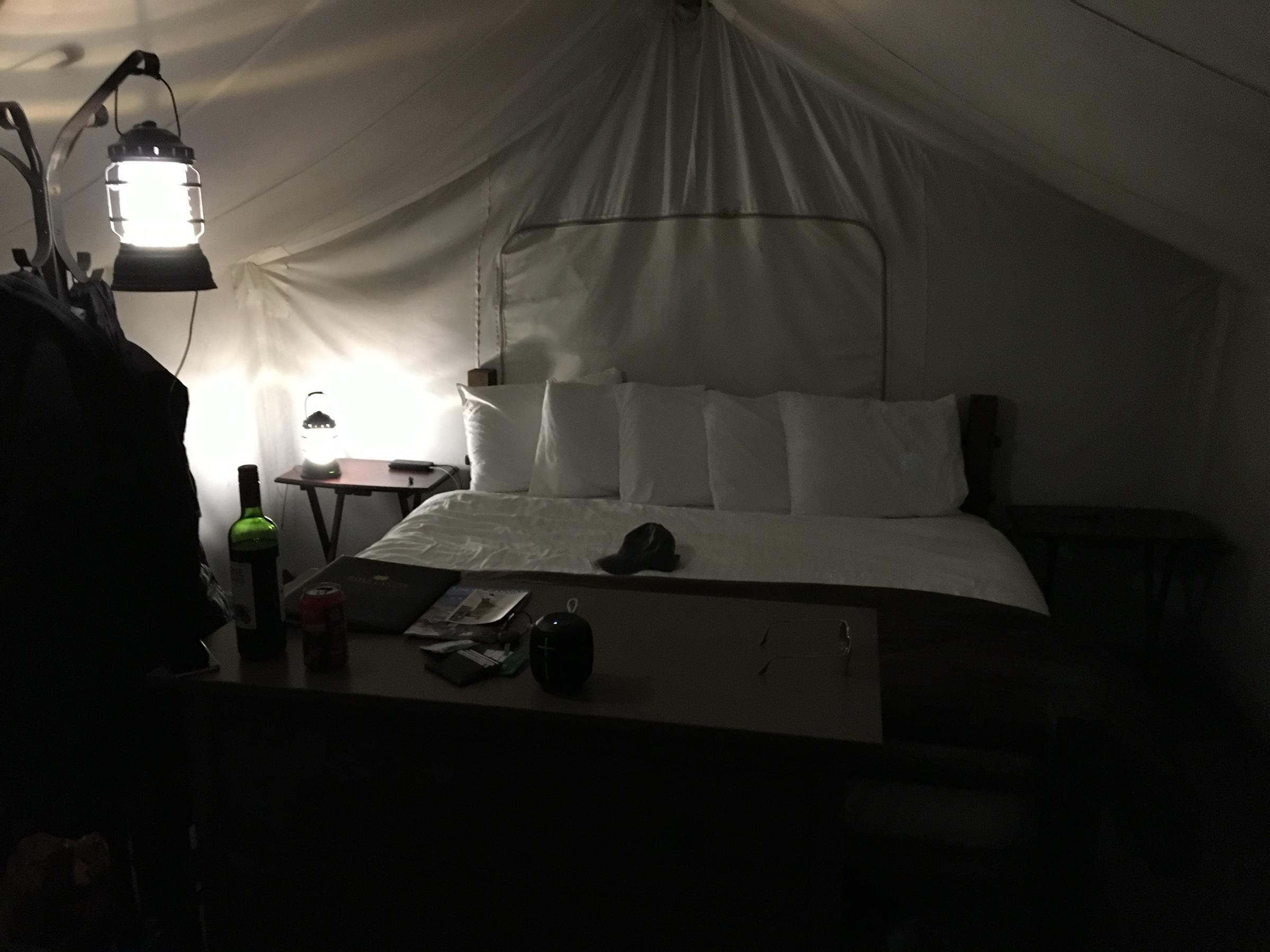 Inside a canvas safari tent at night, a lantern lit beside a large white bed