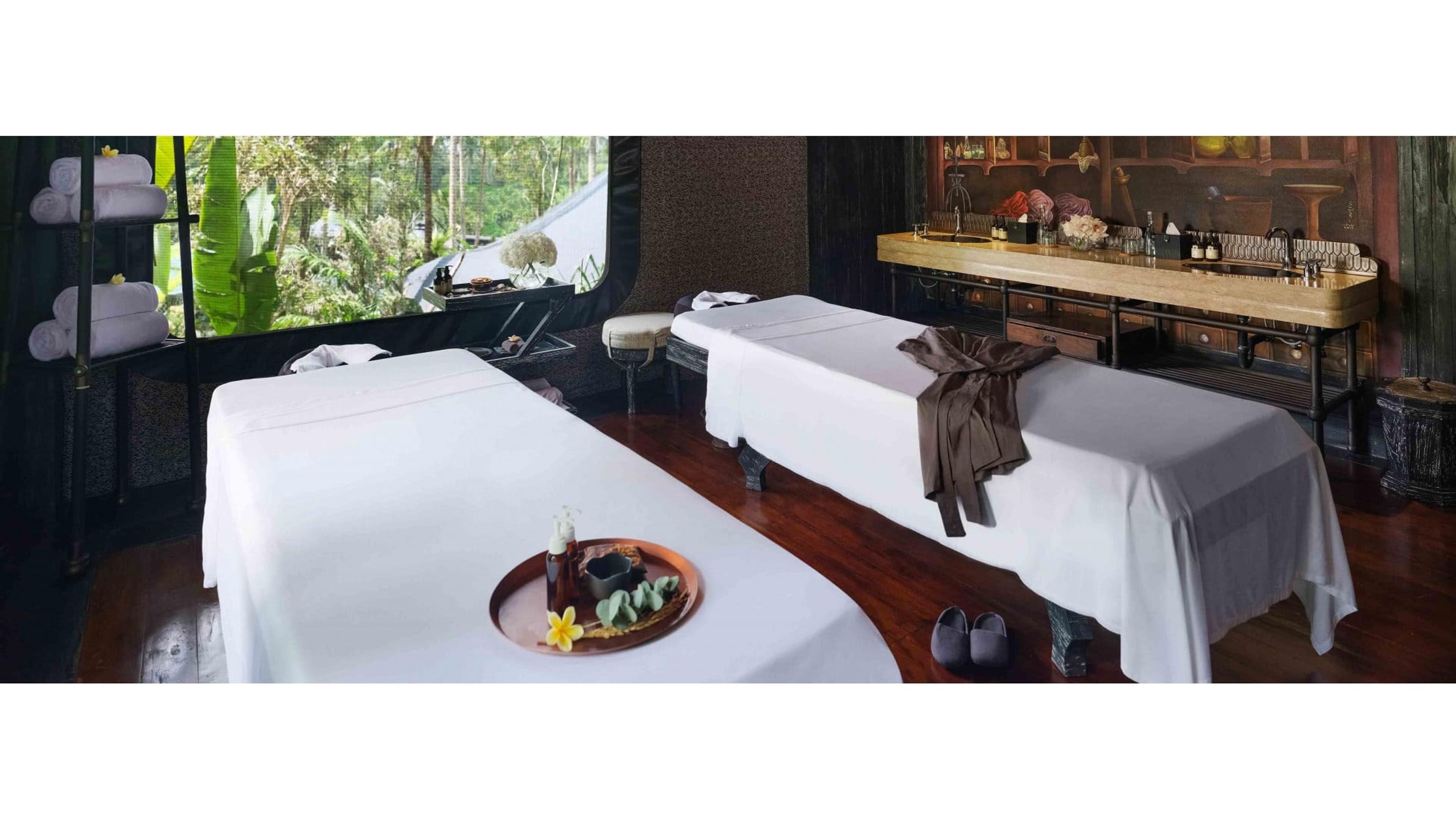 Two massage beds made up with white linens in a spa room, with a window onto the jungle and a drinks counter in the background.