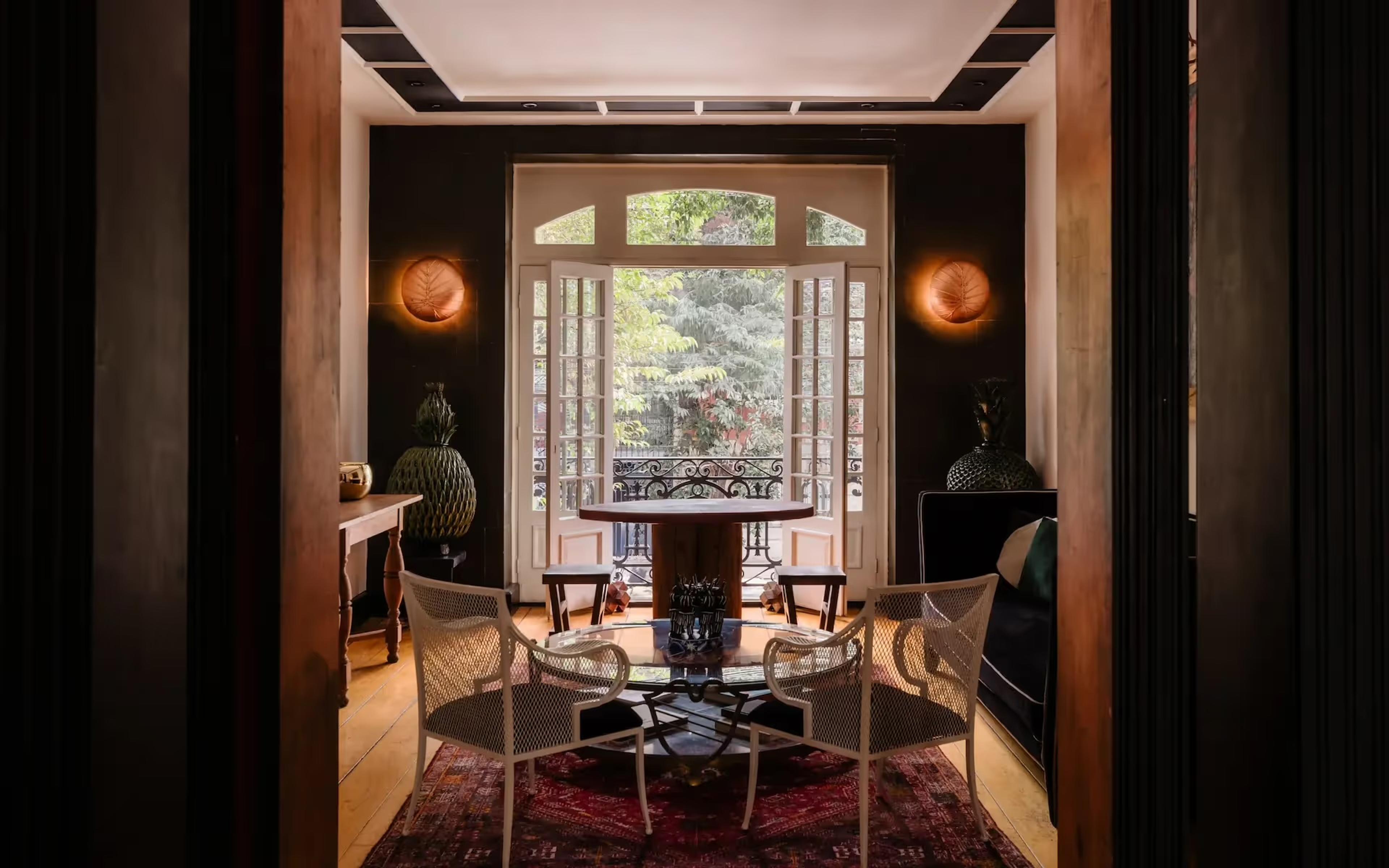 A dark panelled lounge with a round table and cane-back chairs on a patterned rug, French doors open onto a wrought-iron balcony above the street.