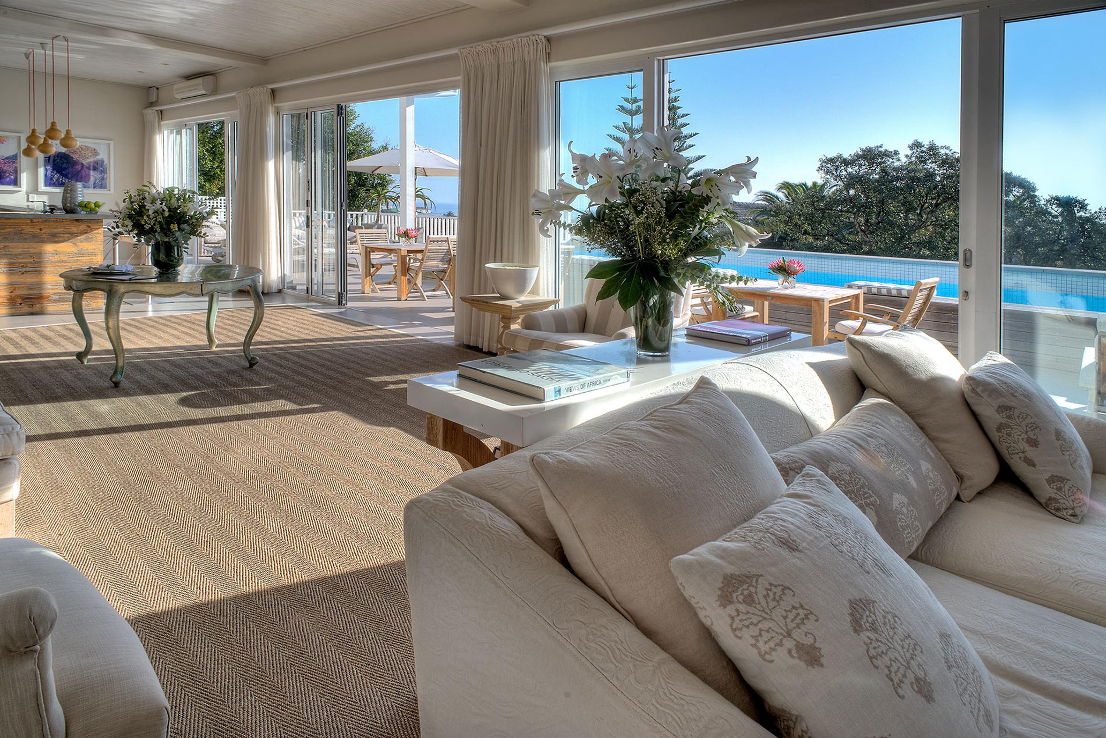 A living room seen through open glass doors, with a pale sofa, a tall vase of lilies on a low table, and a pool beyond the glass.
