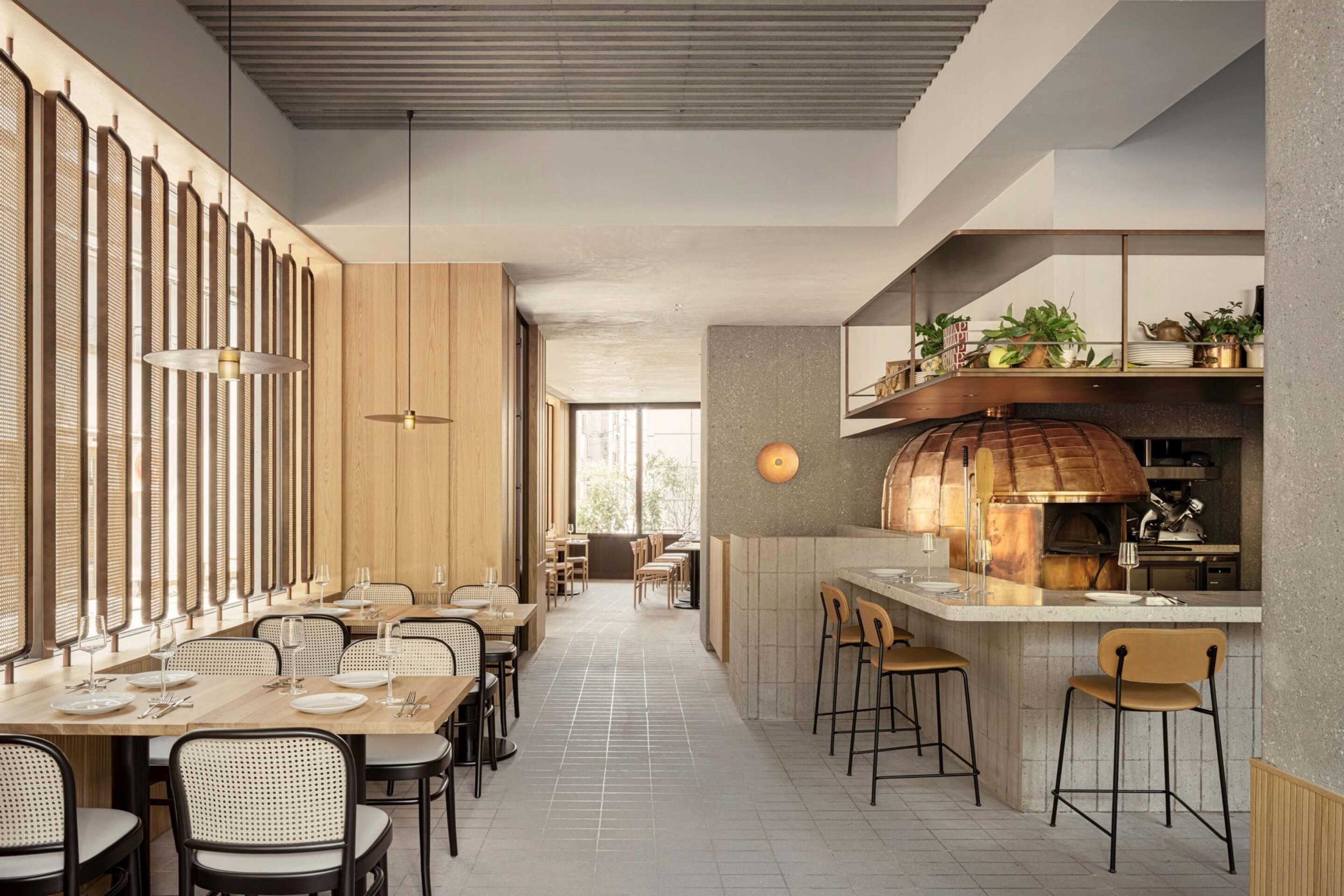 a restaurant dining room with wood-panelled booths and pendant lights leading to a bar with a copper pizza oven and open shelving