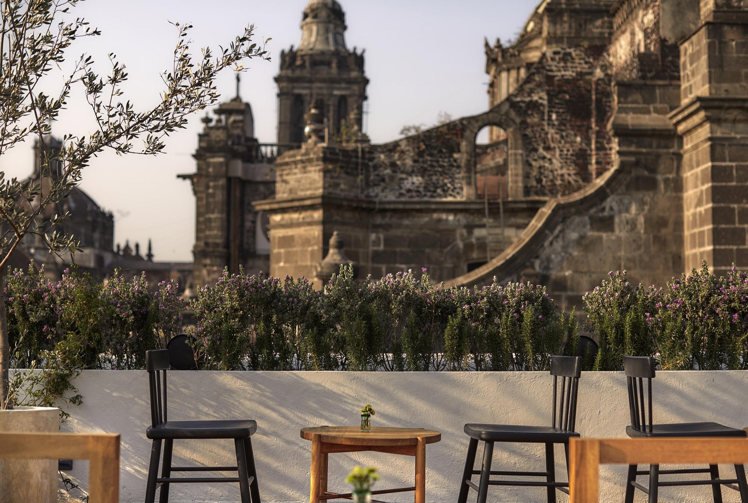 A rooftop terrace with a blurred table and chairs in the foreground, flowering shrubs along a low wall, and a weathered stone church facade behind.