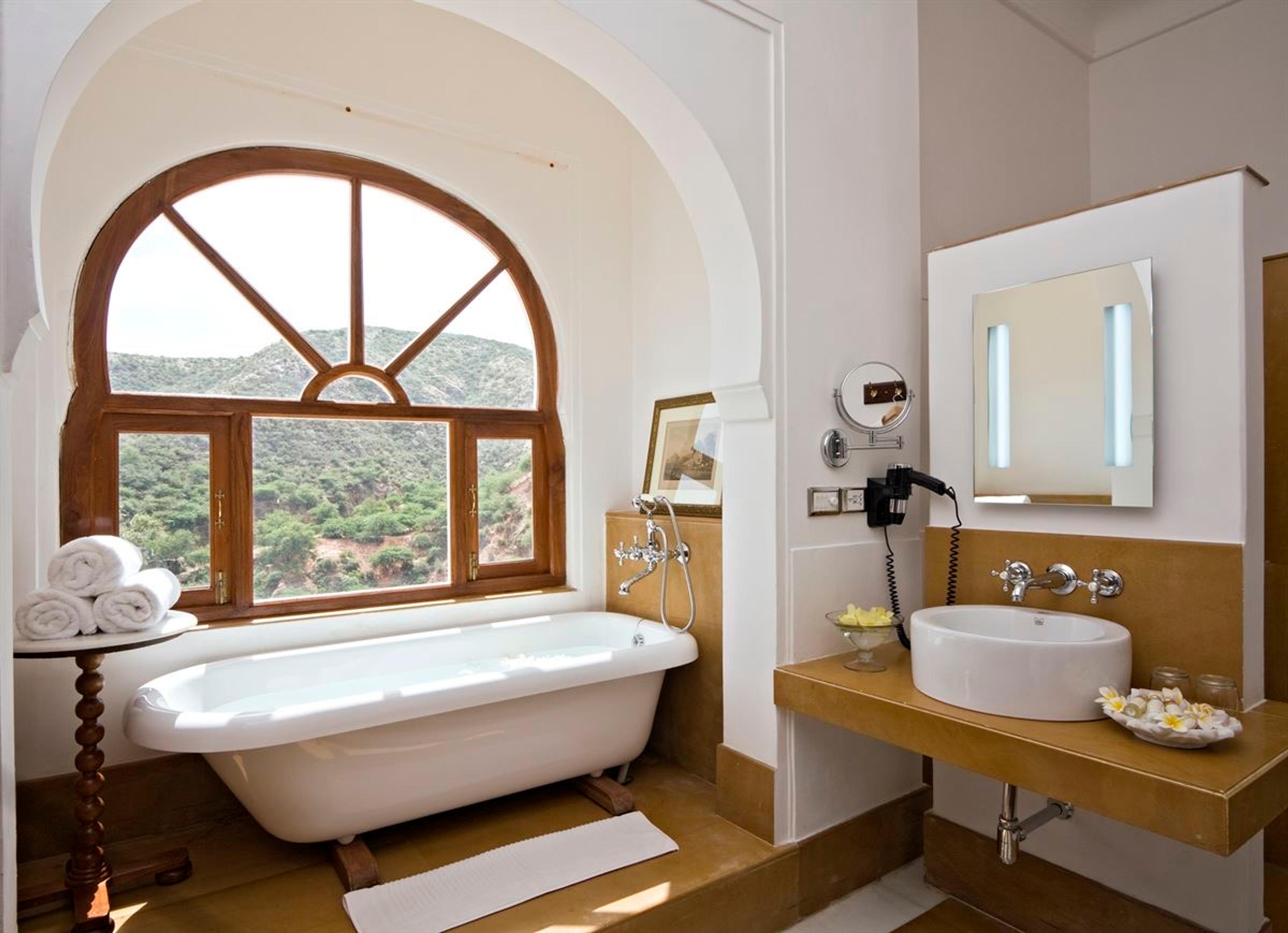 A freestanding bathtub beneath an arched window looking out over wooded hills, with a sink and mirror to the right.