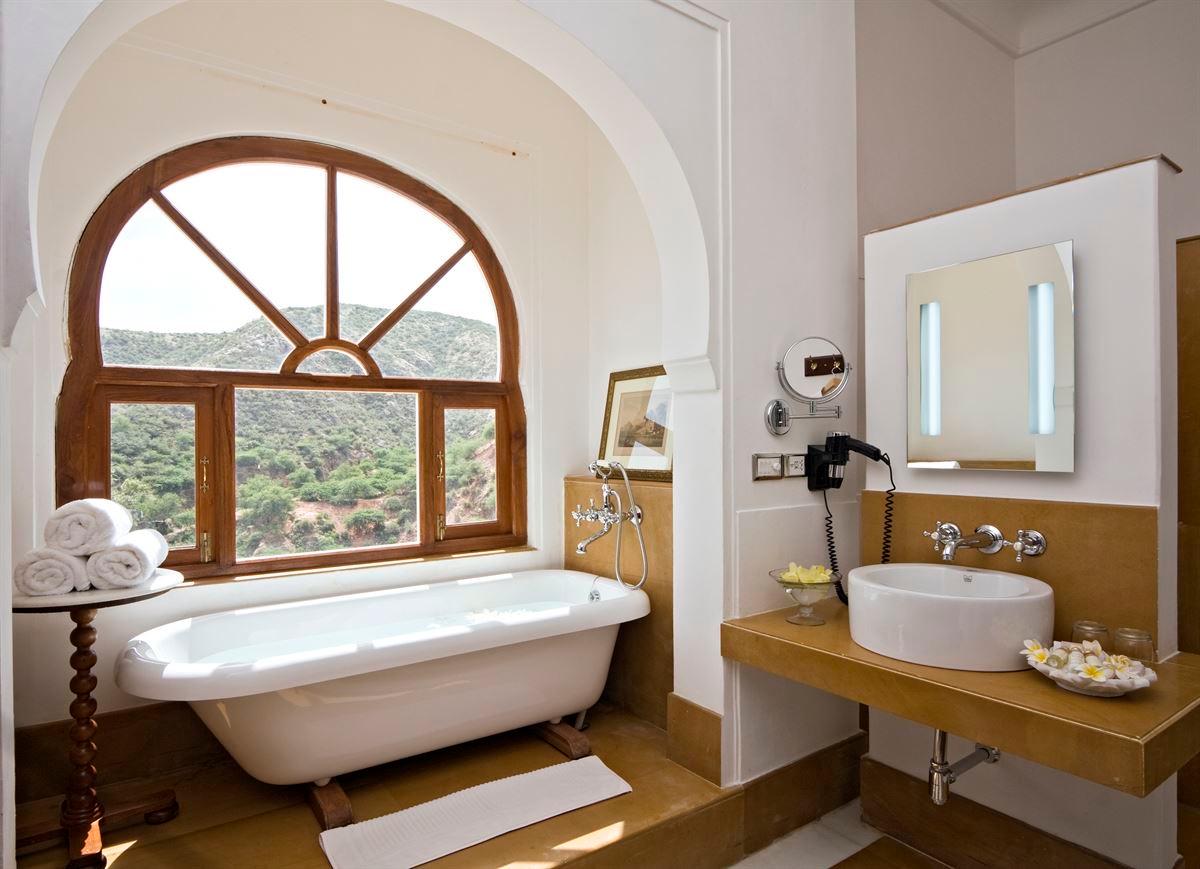 A freestanding bathtub beneath an arched window looking out over wooded hills, with a sink and mirror to the right.