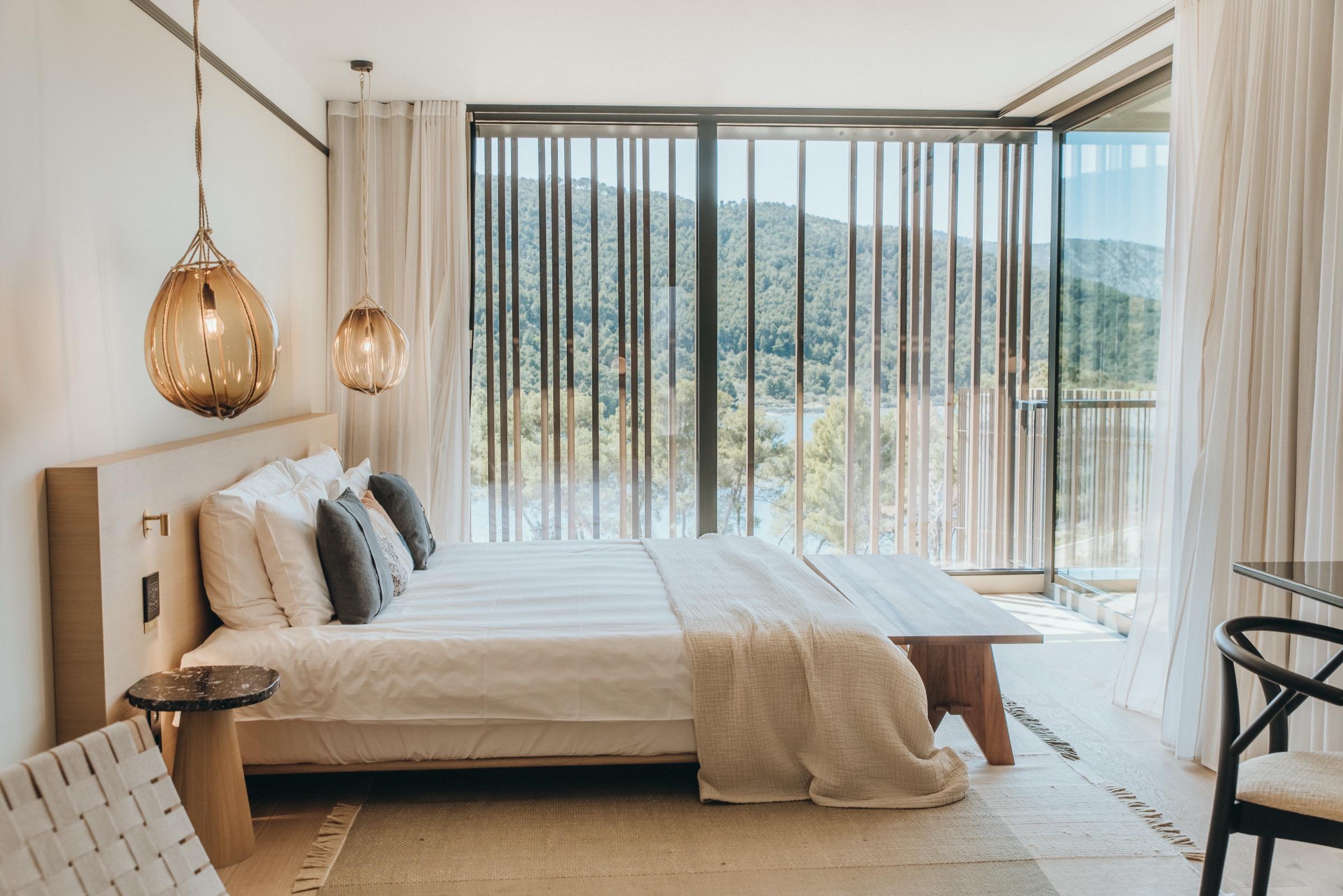 A bedroom with a made bed, two pendant lamps, and floor-to-ceiling slatted glass onto a forested hillside and a stretch of water.