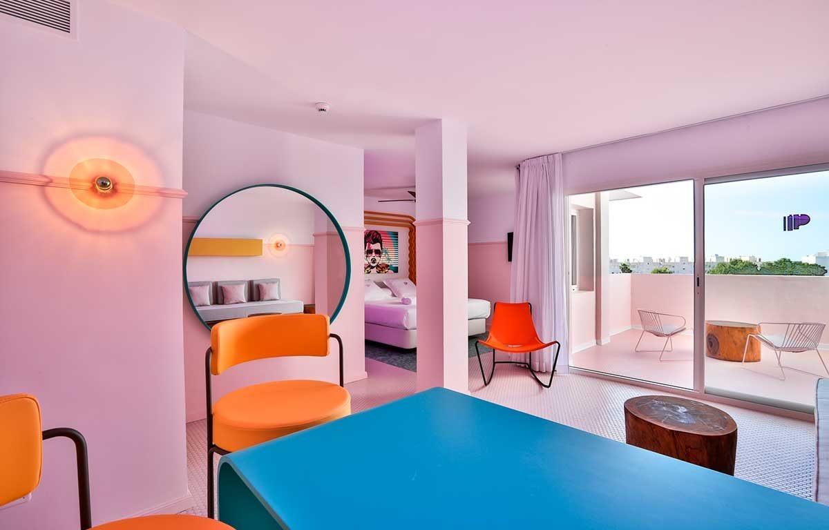 A pink room with a blue table and orange chairs in the foreground, a round mirror on the left reflecting a bed, and sliding doors onto a small terrace with two white chairs and a tree-trunk table.