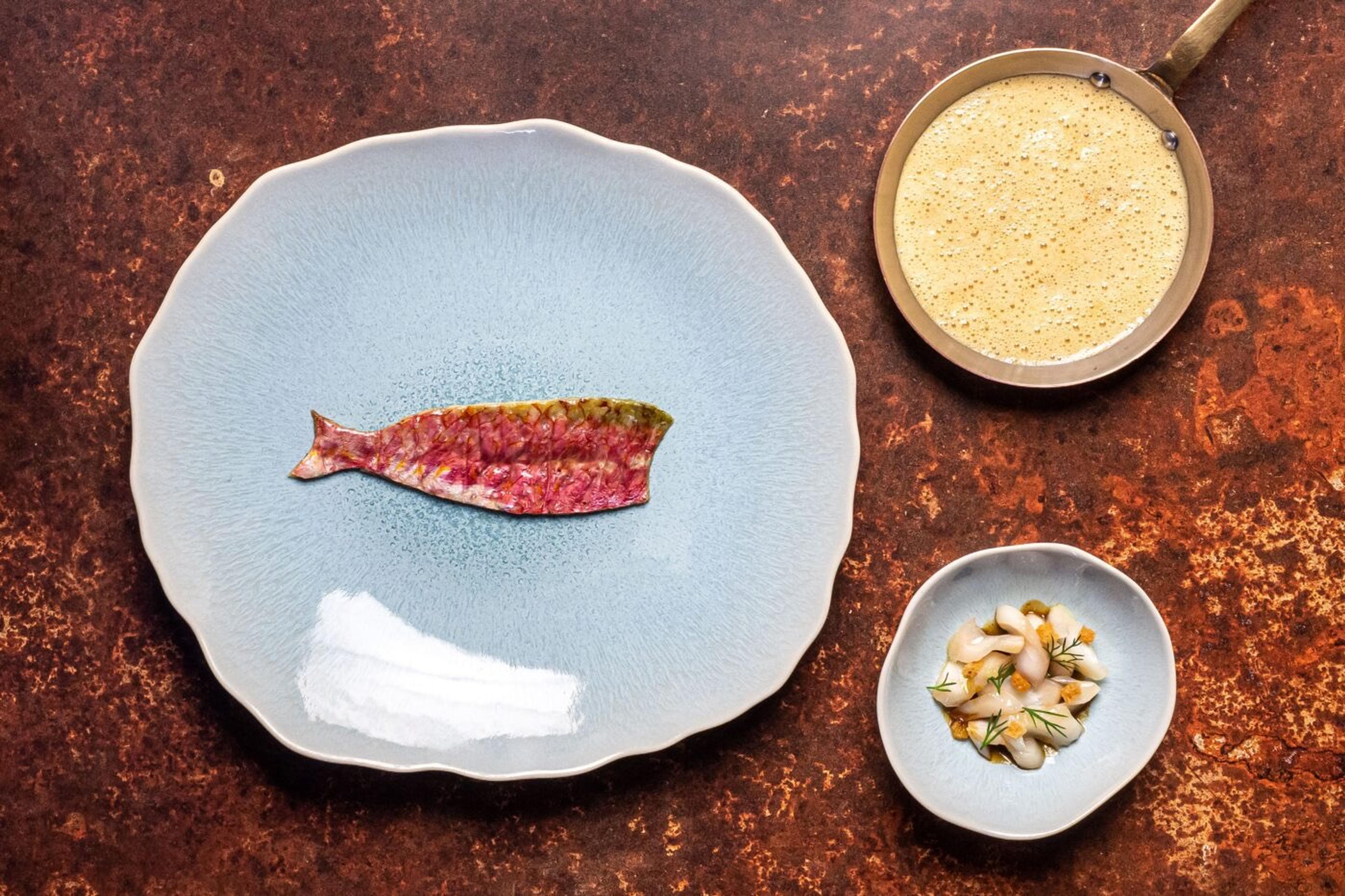 An overhead shot of a plated red mullet fillet on a pale blue plate, beside a small bowl of shellfish and a pan of foamed sauce.