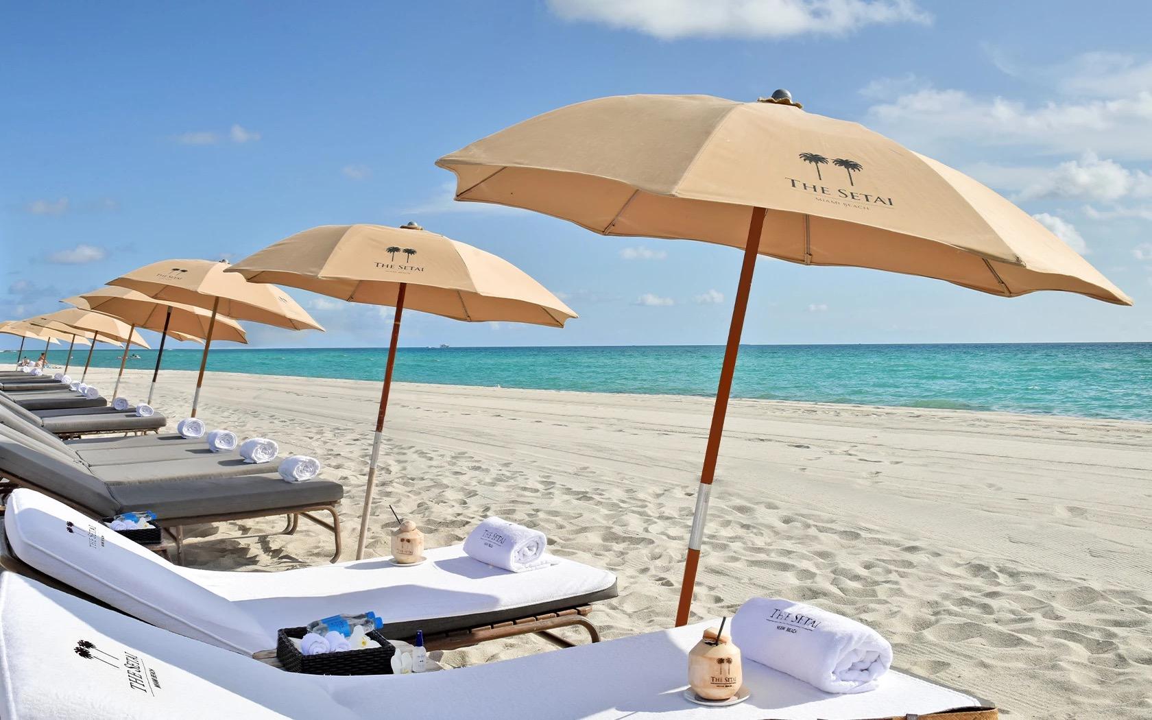 A row of cream sun loungers and umbrellas branded with The Setai's palm-tree logo line a beach beside turquoise water.