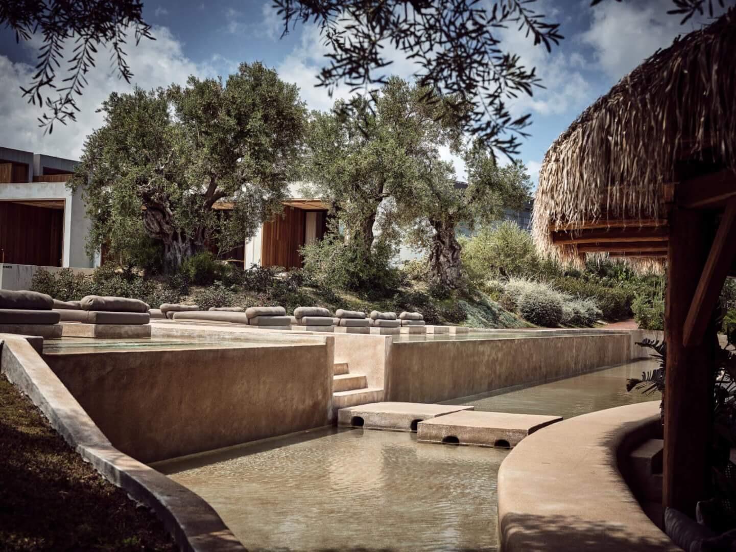 A stepped water channel runs past olive trees and a thatched pavilion, with low stone walls and cushioned seating alongside.