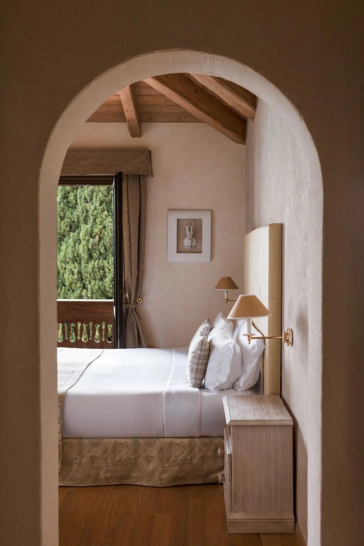 A stone archway frames a bed with a patterned throw under a wood-beamed ceiling, a window open to greenery beside it.