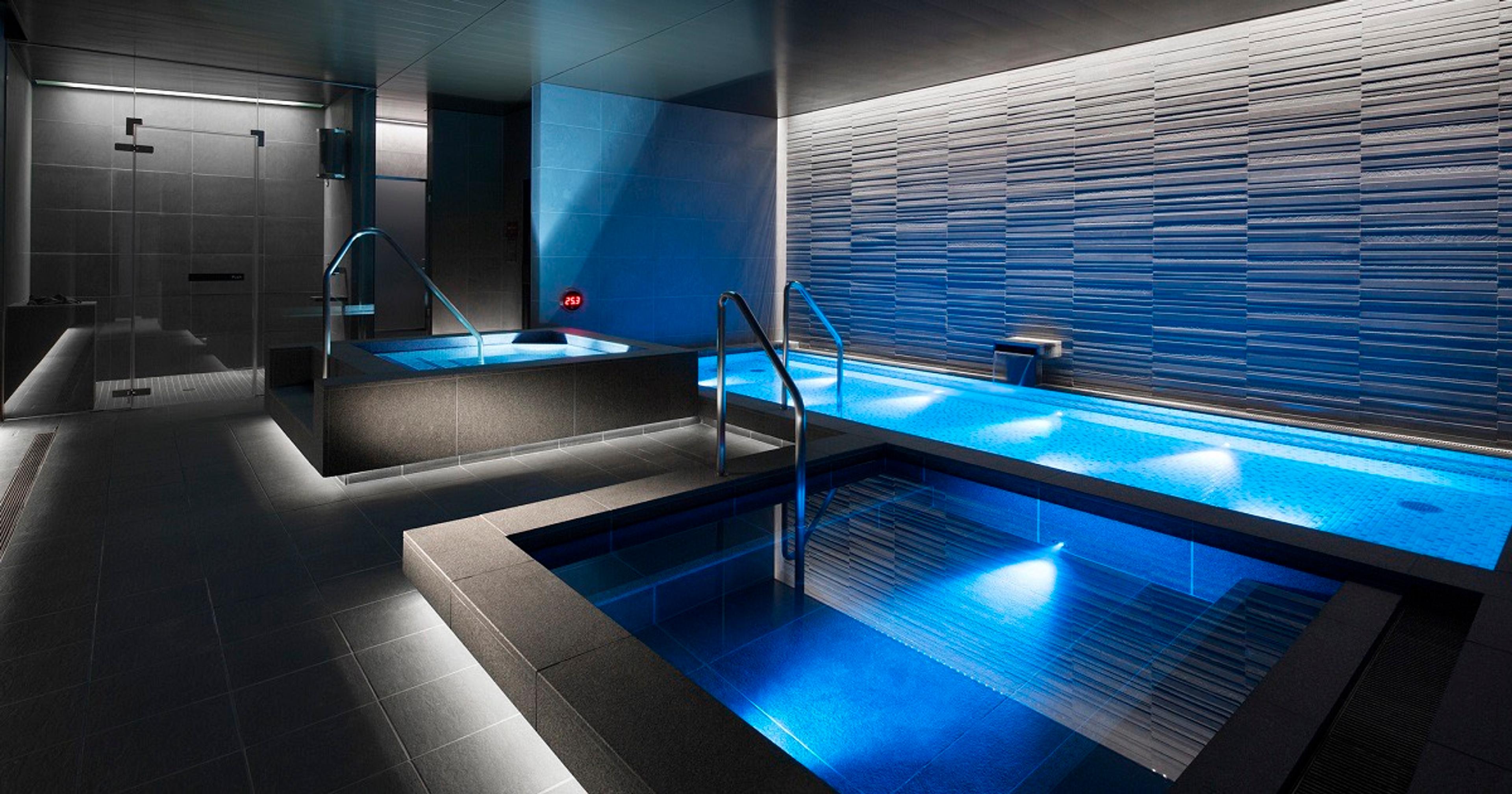 An indoor spa with a dimly lit blue-tiled plunge pool, steps and handrails, and a glass shower enclosure to one side.