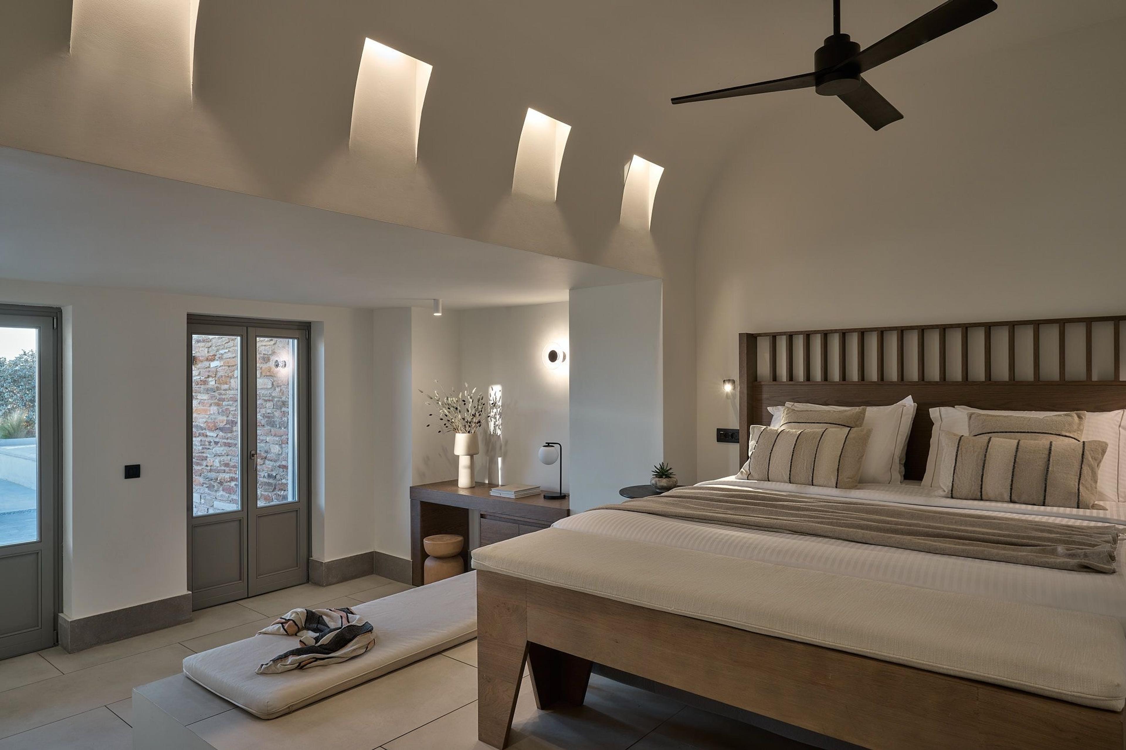 A bedroom with a timber-framed bed under lit arched niches in a vaulted ceiling, a ceiling fan, and a desk with a lamp against the far wall.