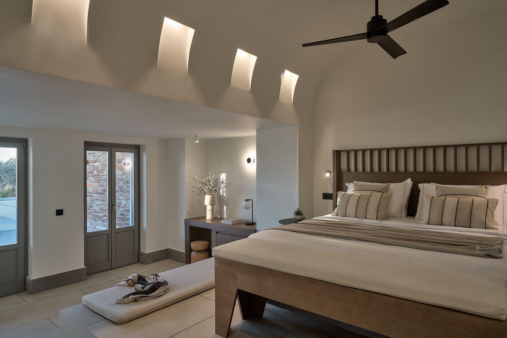A bedroom with a timber-framed bed under lit arched niches in a vaulted ceiling, a ceiling fan, and a desk with a lamp against the far wall.