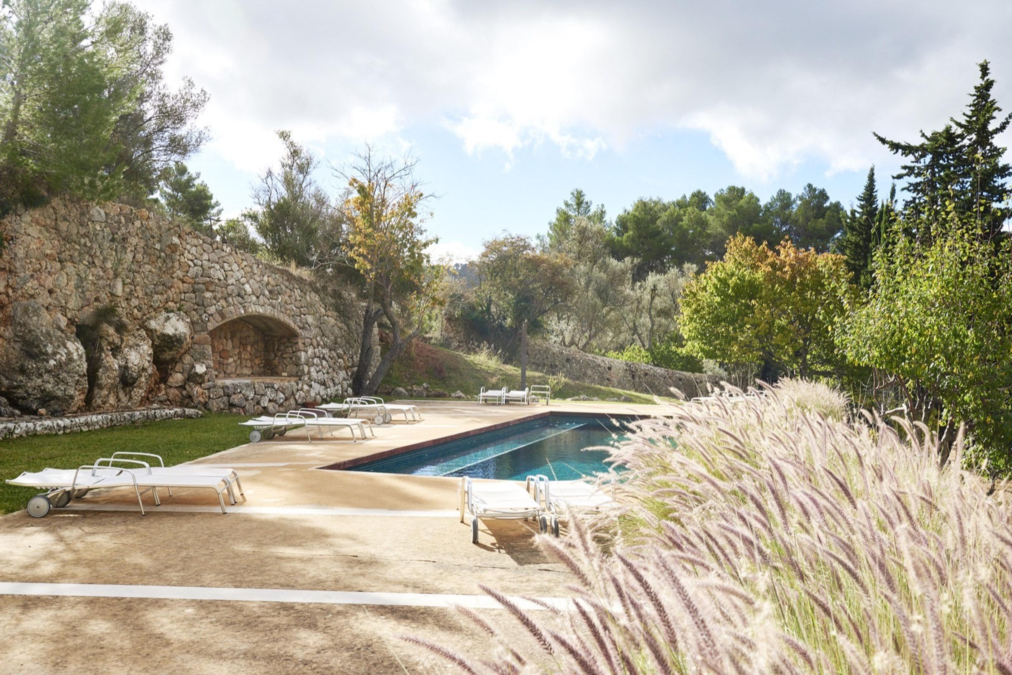 A swimming pool beside a rustic stone wall with an arched grotto niche, loungers on the terrace around it and ornamental grasses in the foreground.