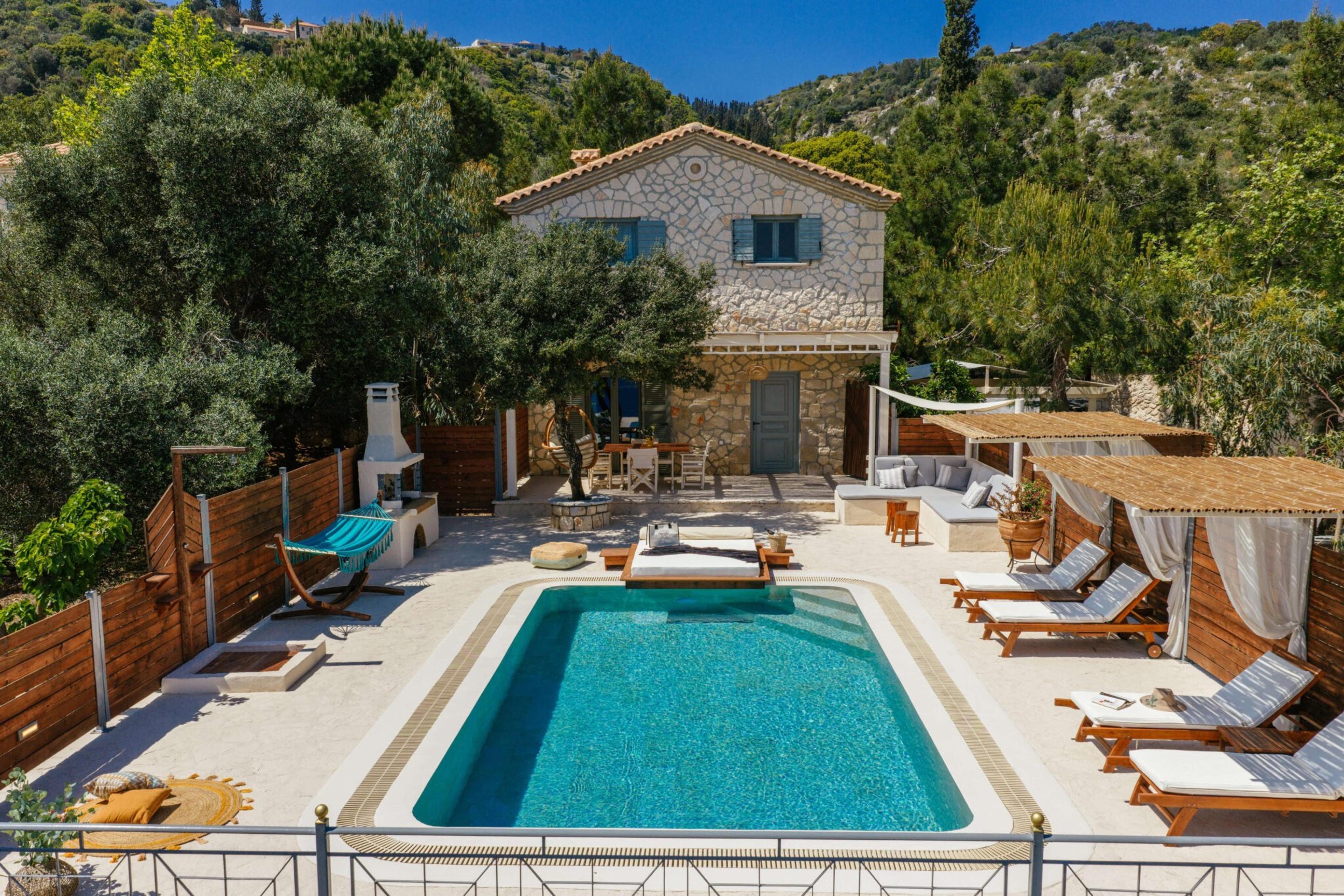 a stone villa with blue shutters seen across a pool and sun loungers, hills and trees rising behind it