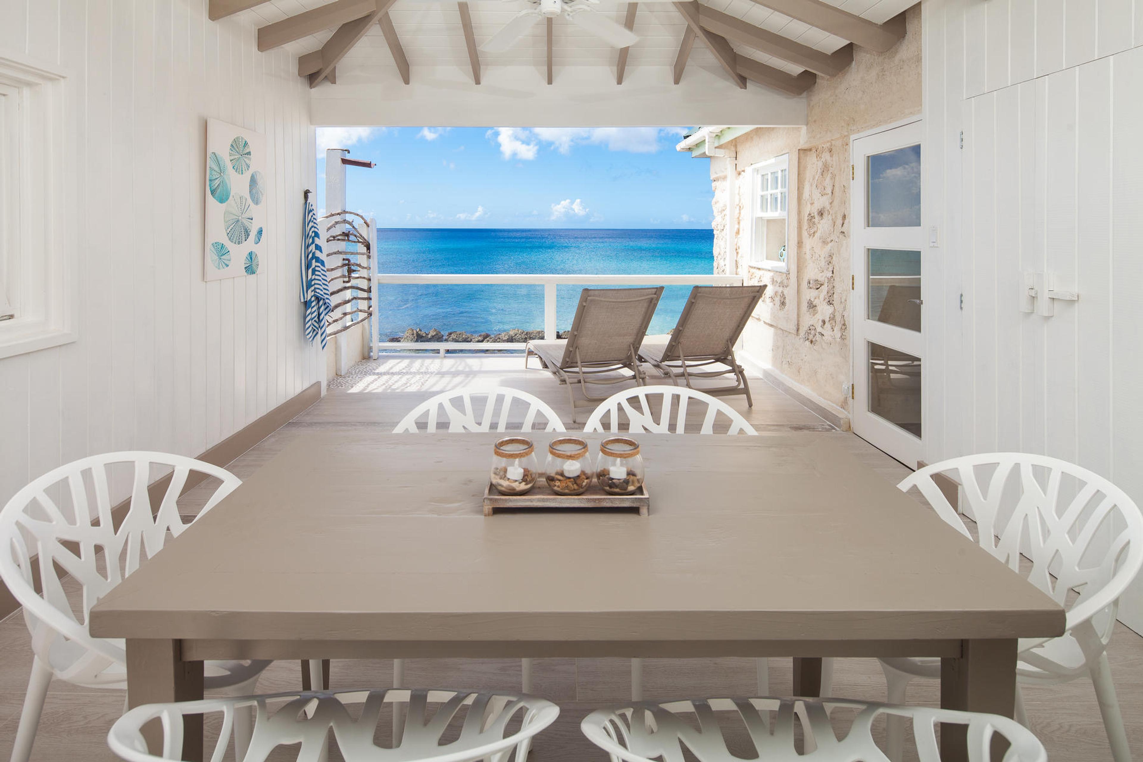 A covered terrace with a long table set for eight and white chairs, sun loungers facing the open sea beyond the railing.
