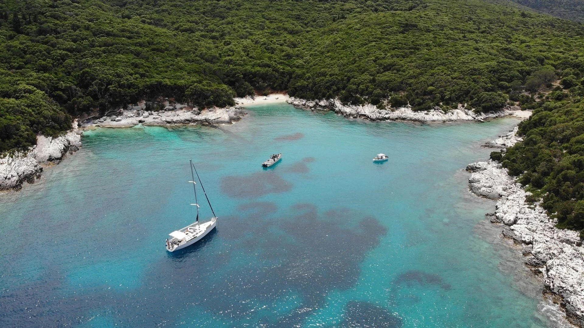 An aerial view of a secluded turquoise cove ringed by pine-covered cliffs, a sailing boat and two small boats anchored in the bay.