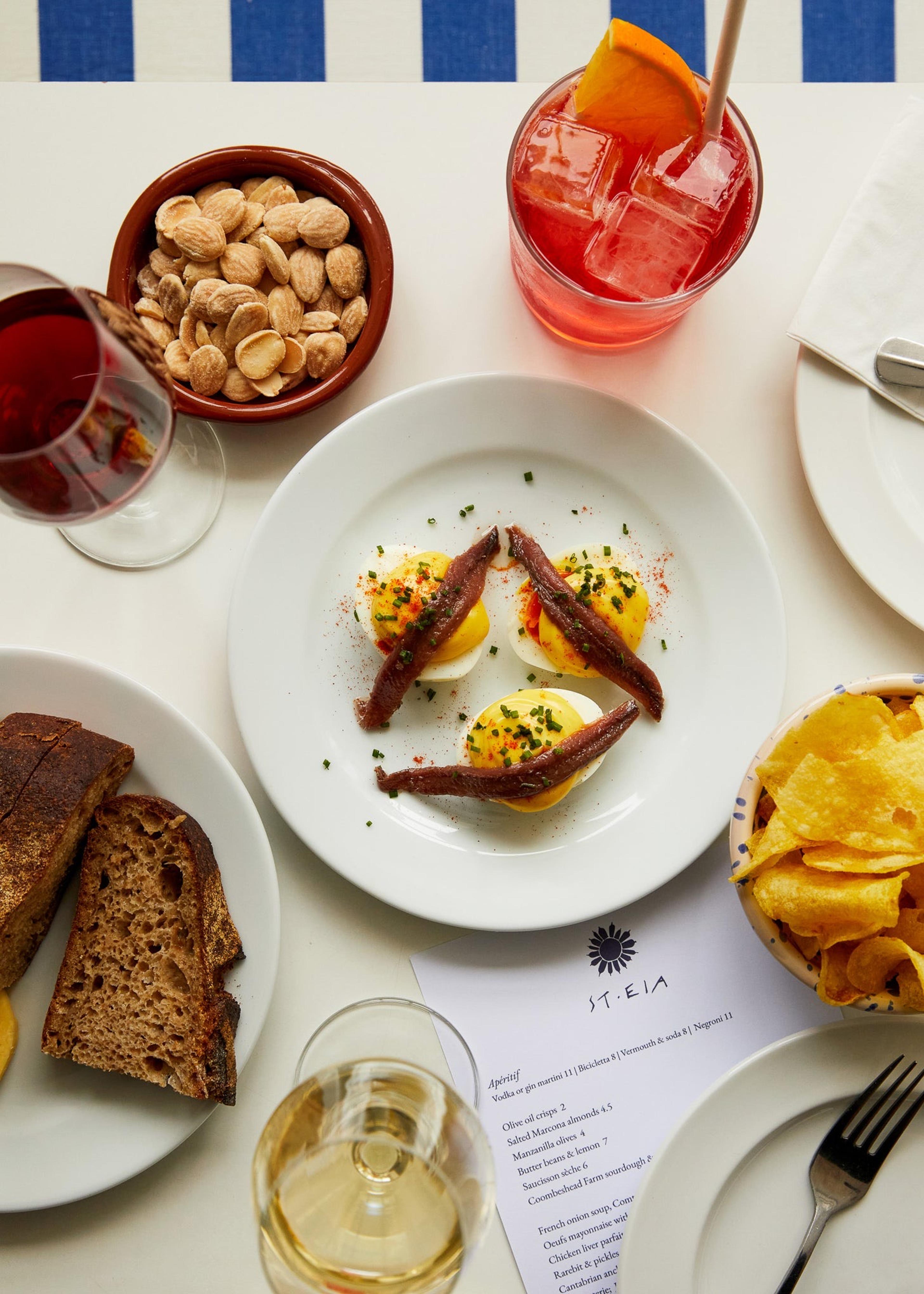 Small plates on a white table: anchovies over egg, bread, olives and a glass of red wine.