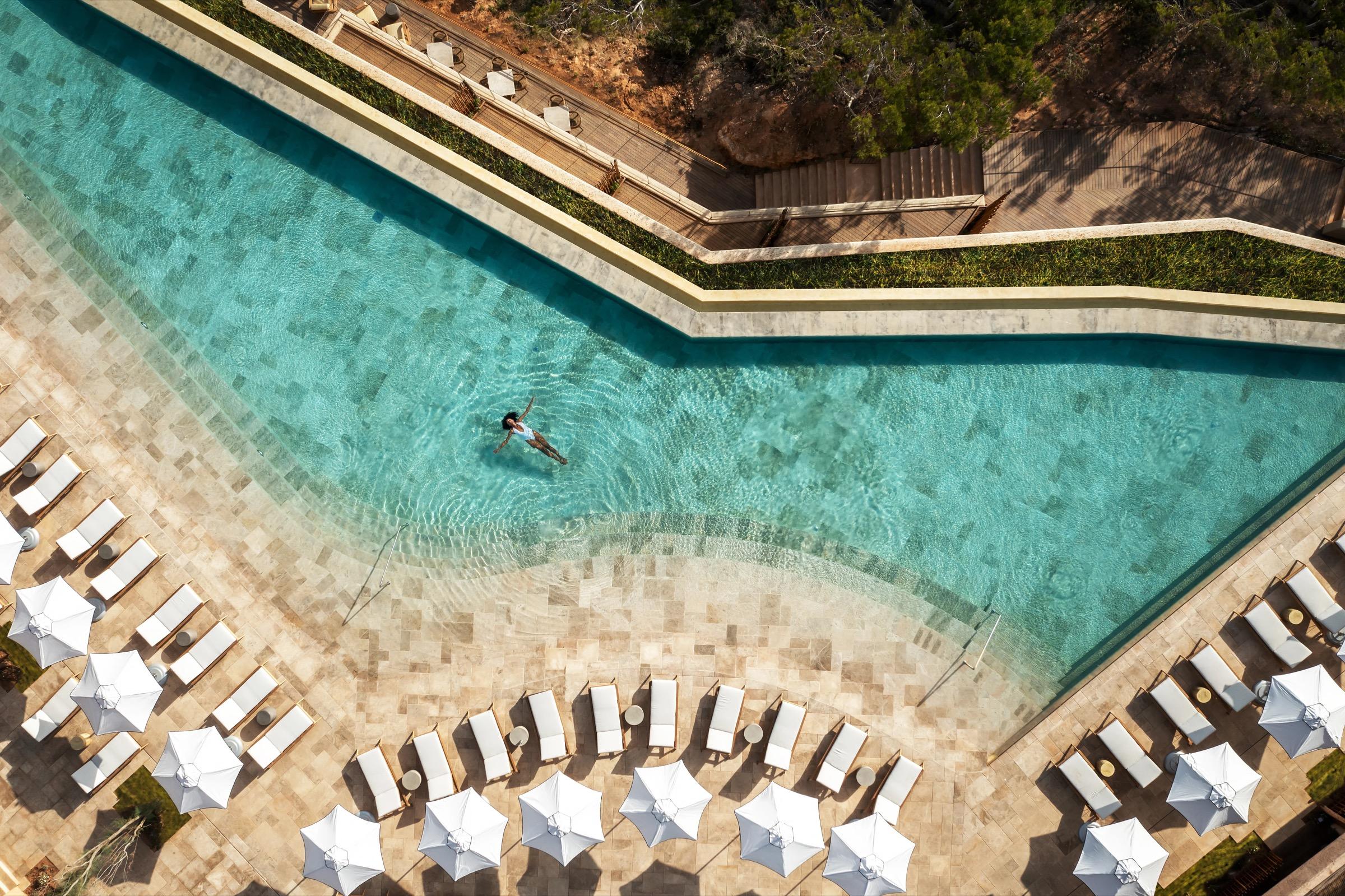 Aerial view of a long pool on a stone terrace, with a swimmer in the middle of the water, white umbrellas and loungers around the edge, and the sea beyond.