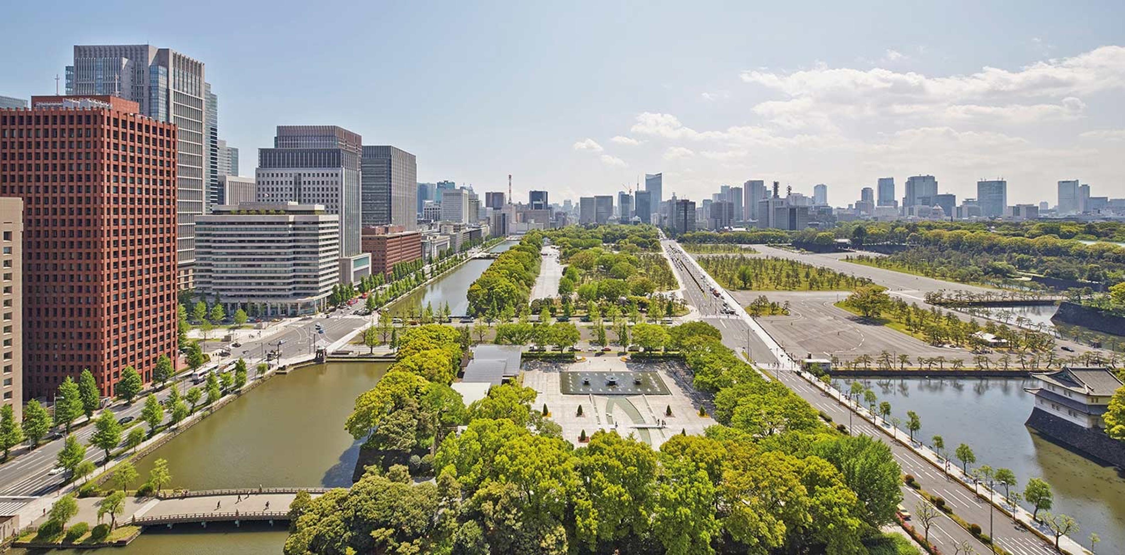 An aerial view over a riverside park and moat, with the Tokyo skyline stretching behind it.
