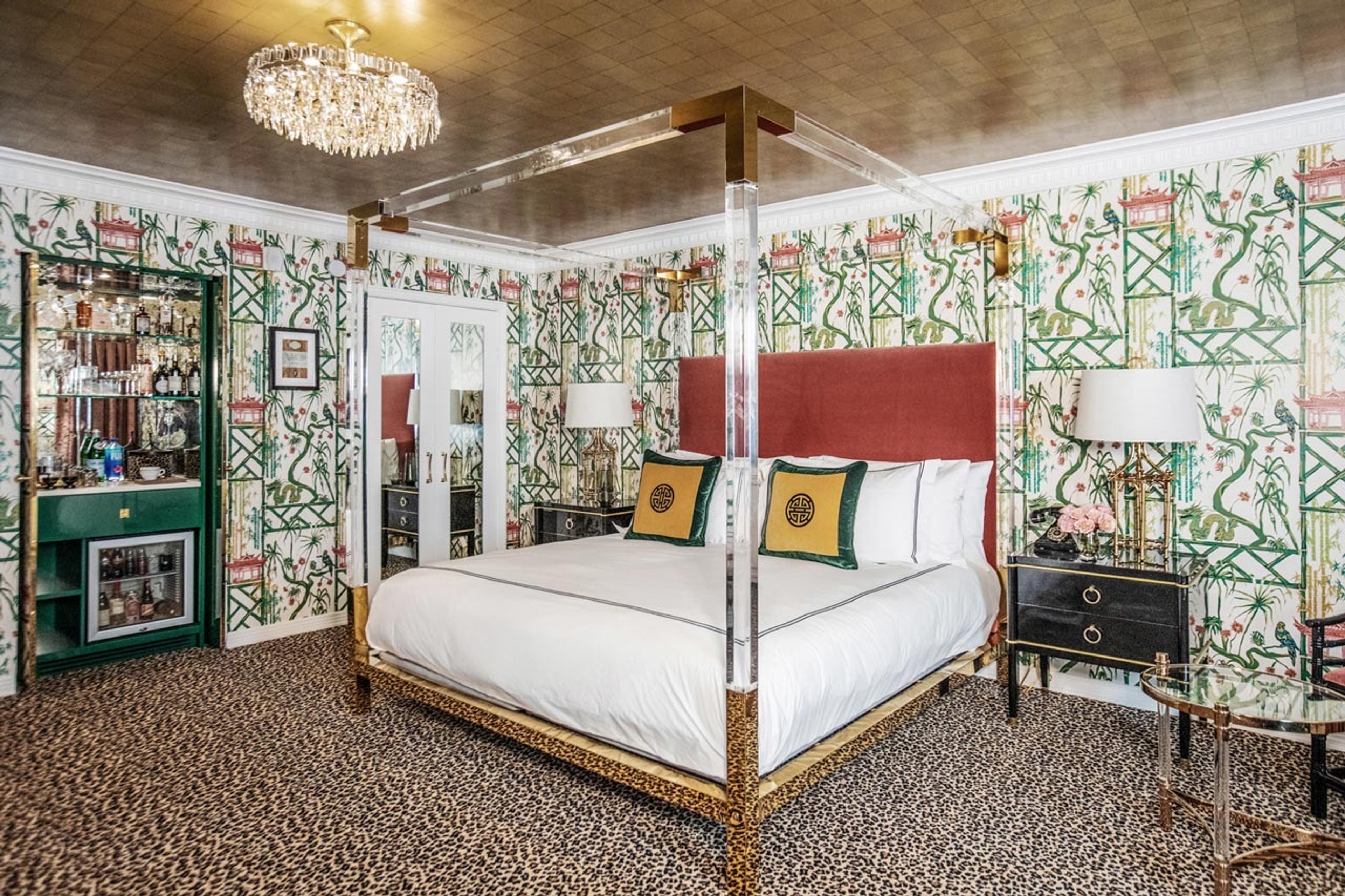 A bedroom with a lucite four poster bed on a leopard print carpet, a dark red headboard, green bamboo and pagoda wallpaper, a crystal chandelier, and a mirrored drinks cabinet in the corner.