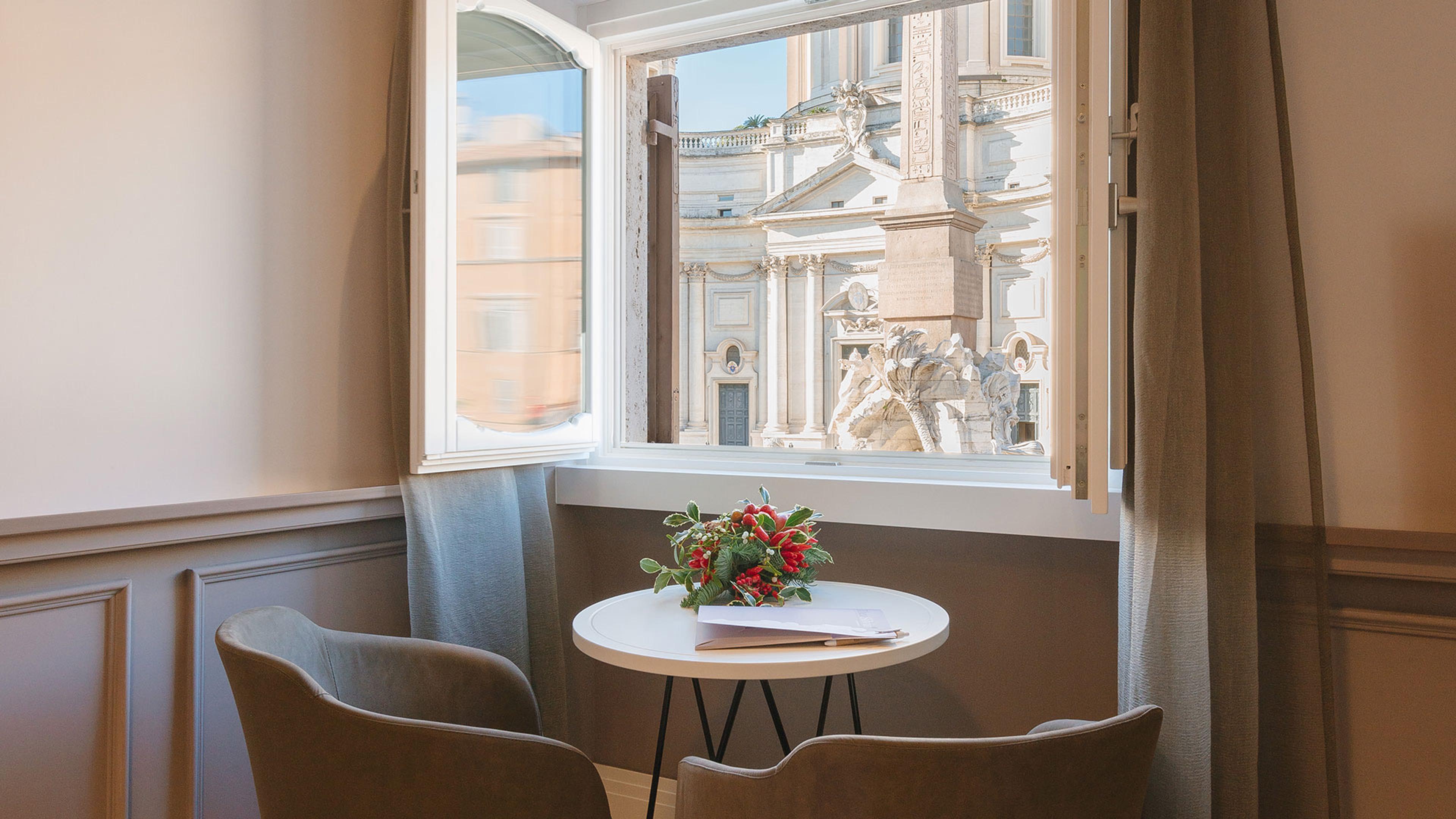 A view through an open window onto a sunlit piazza, with an obelisk, a fountain and a baroque church facade across the square.
