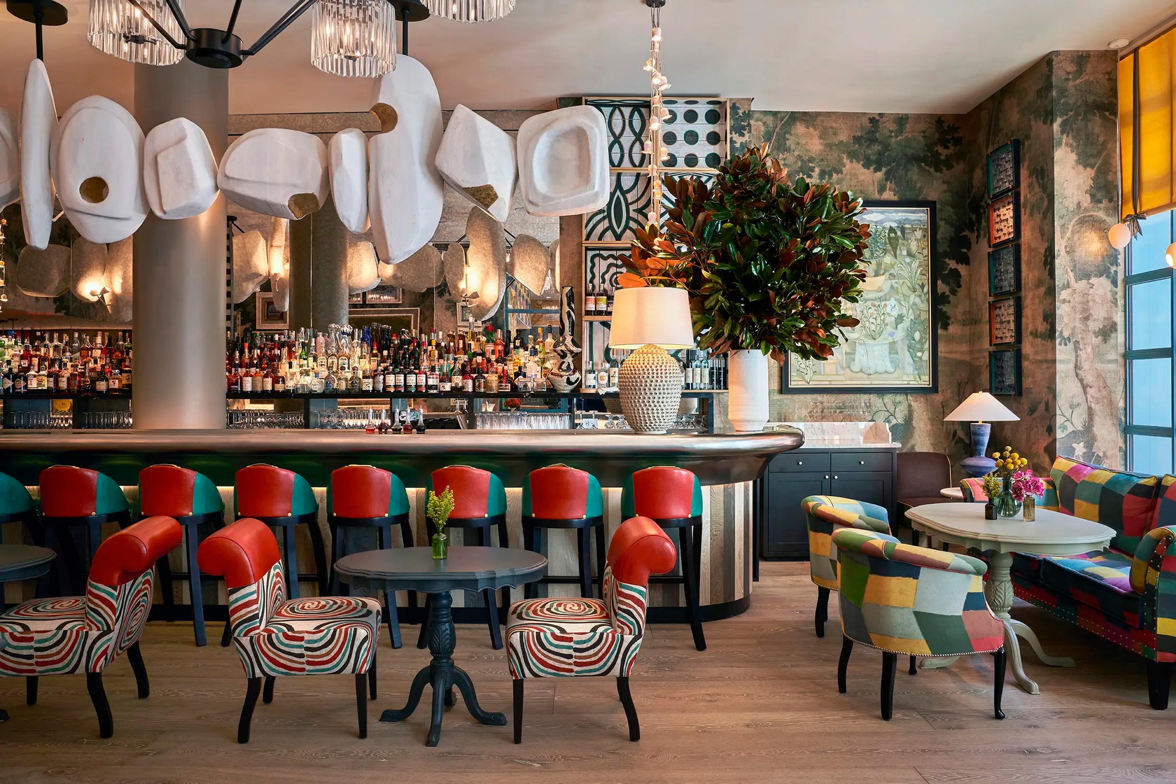 A hotel bar with red leather stools, patterned armchairs around low tables, and a large magnolia branch in a vase on the counter.