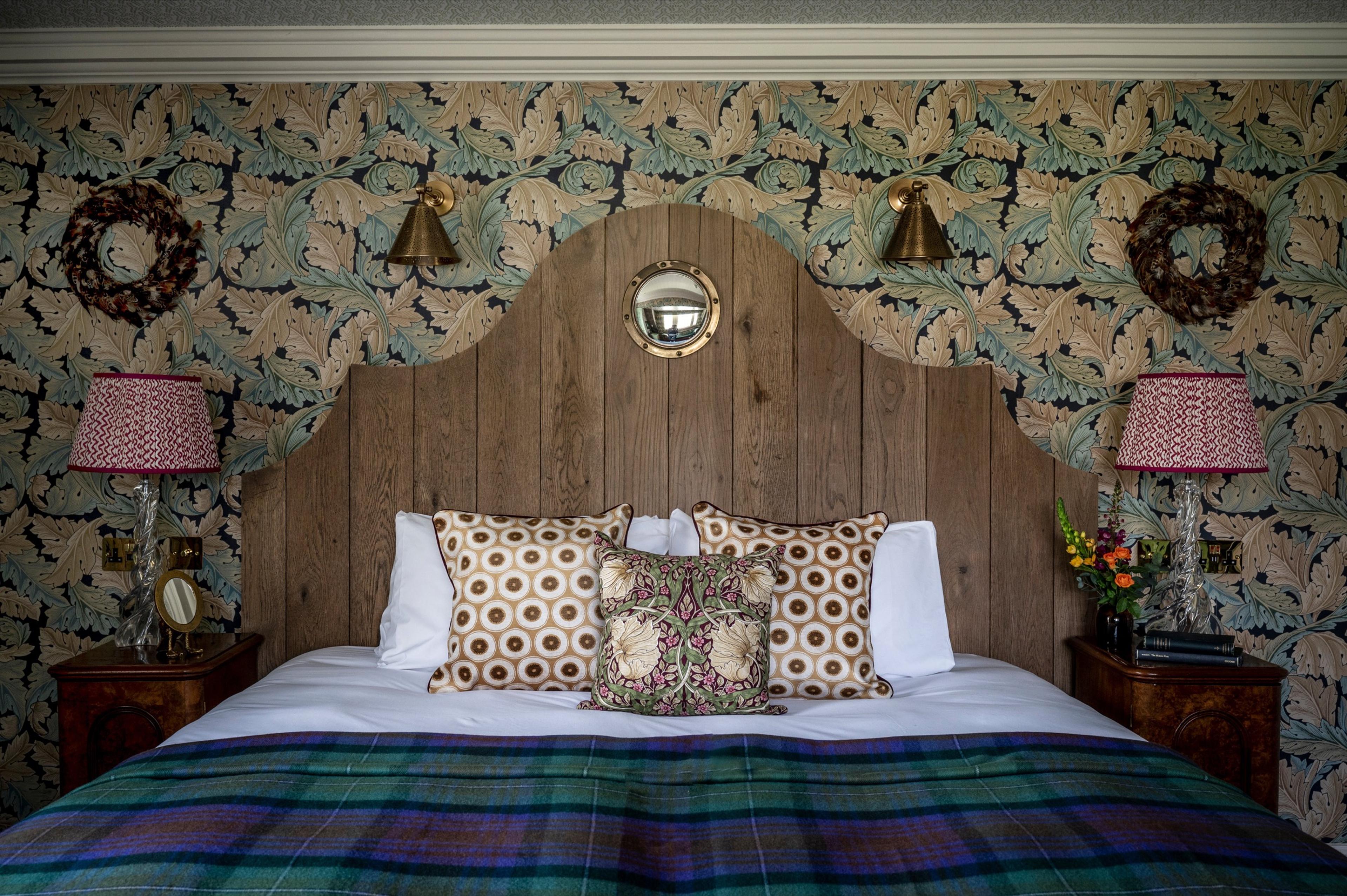 A bed with a carved wooden headboard and tartan blanket, flanked by two lamps, in a room with green leaf-patterned wallpaper