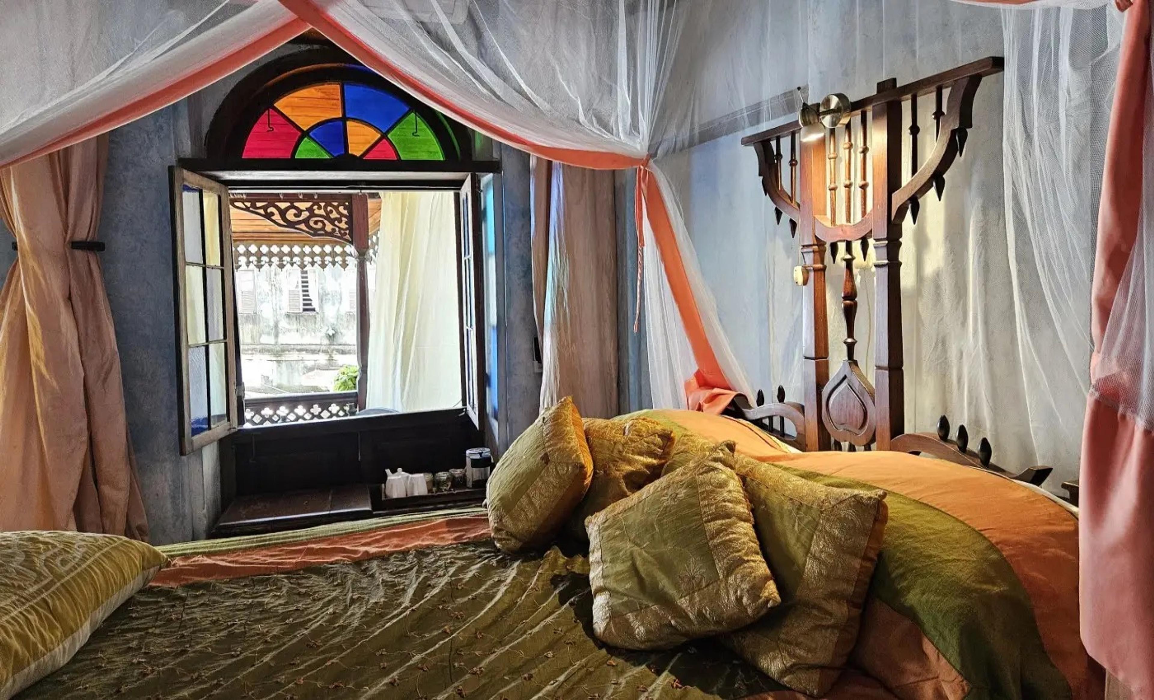 a bed with olive and gold silk pillows under a mosquito net canopy, beside an open stained-glass window looking out over rooftops