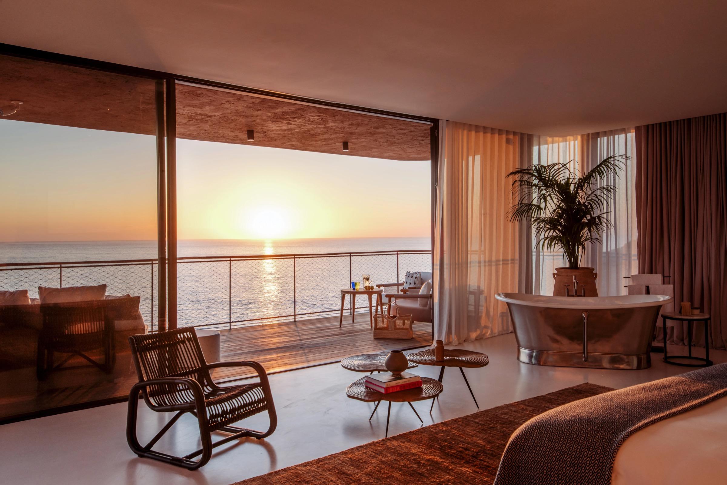 A bedroom at sunset with a made bed at the bottom right, a freestanding bath beside a palm, and a rattan chair and low tables in front of glass doors open onto a balcony over the sea.