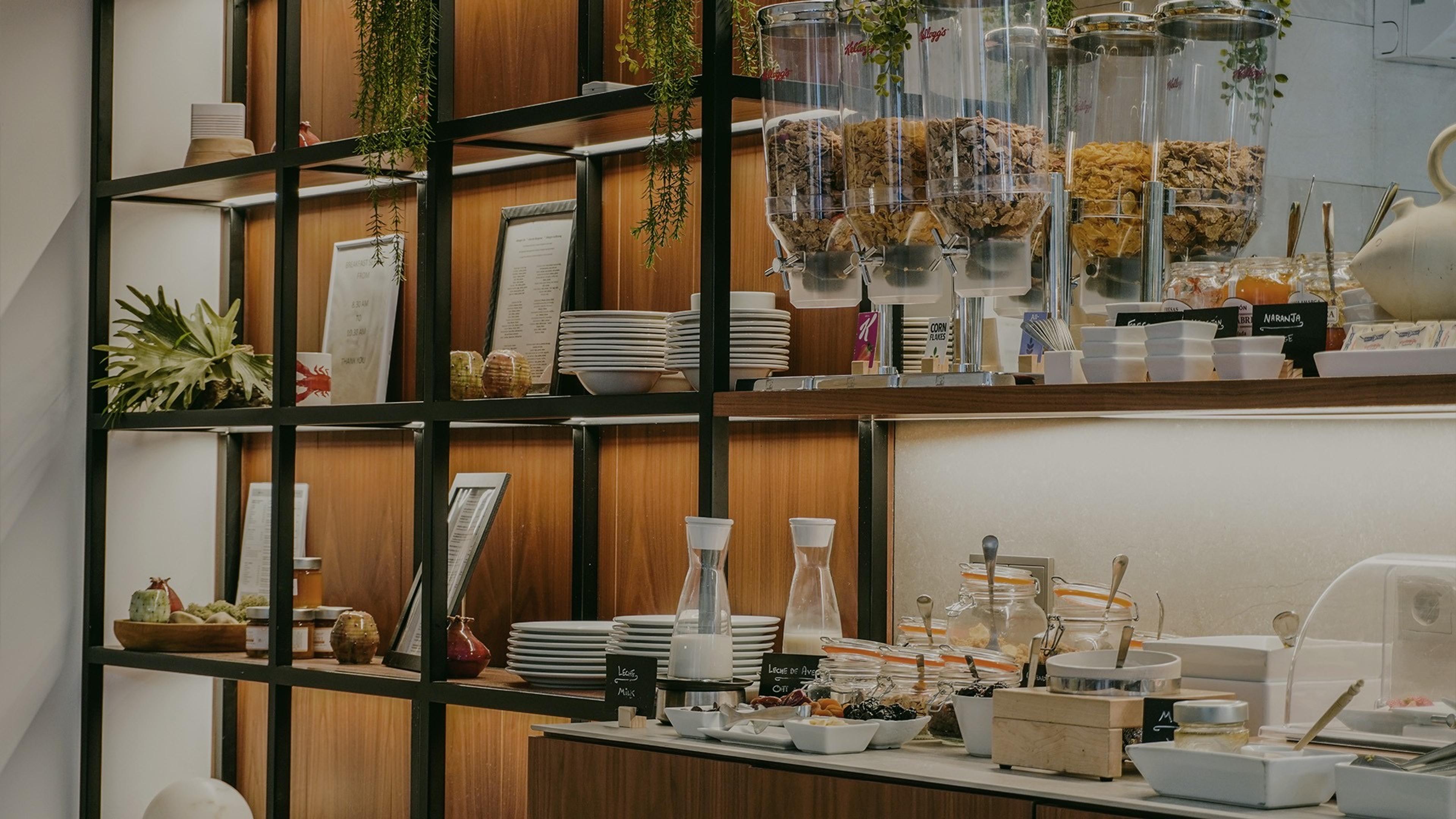 A breakfast counter with cereal dispensers, stacked bowls and plates, jars of nuts and dried fruit, small labelled pots, and a jug, with hanging plants above.
