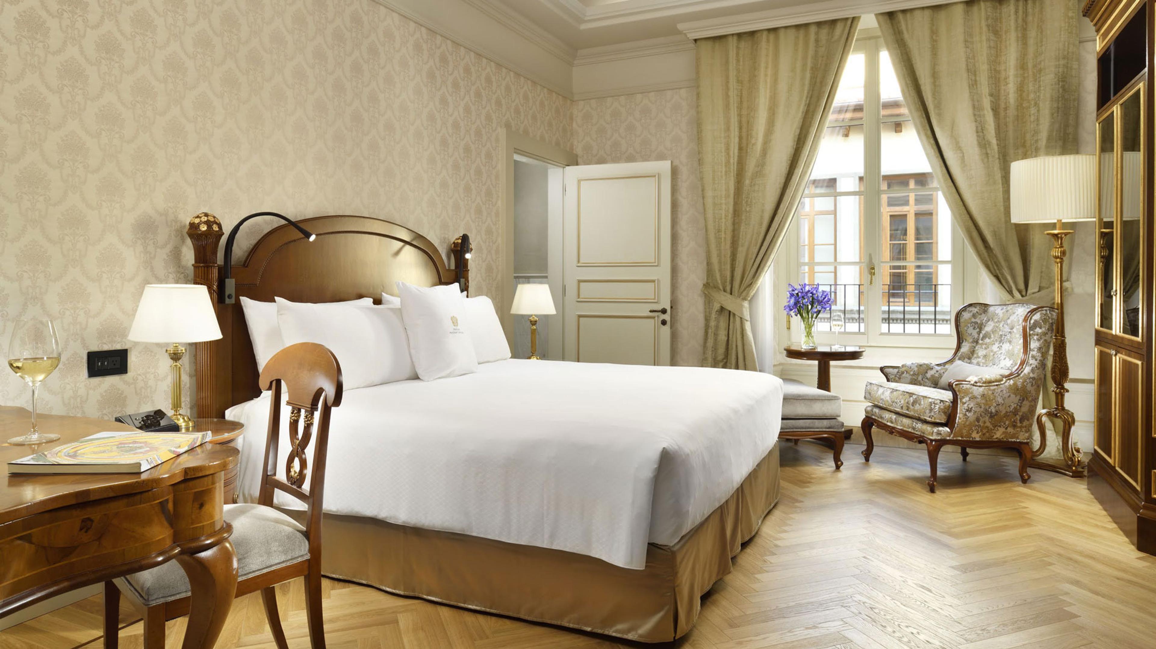 a bedroom with a carved wood bed and white linens, a herringbone parquet floor, a writing desk and a window open to the street