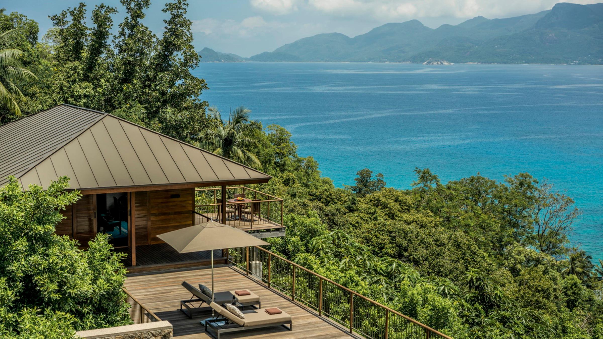 A villa with a pitched roof and a timber deck holding two sun loungers, surrounded by jungle with the sea and mountains beyond.