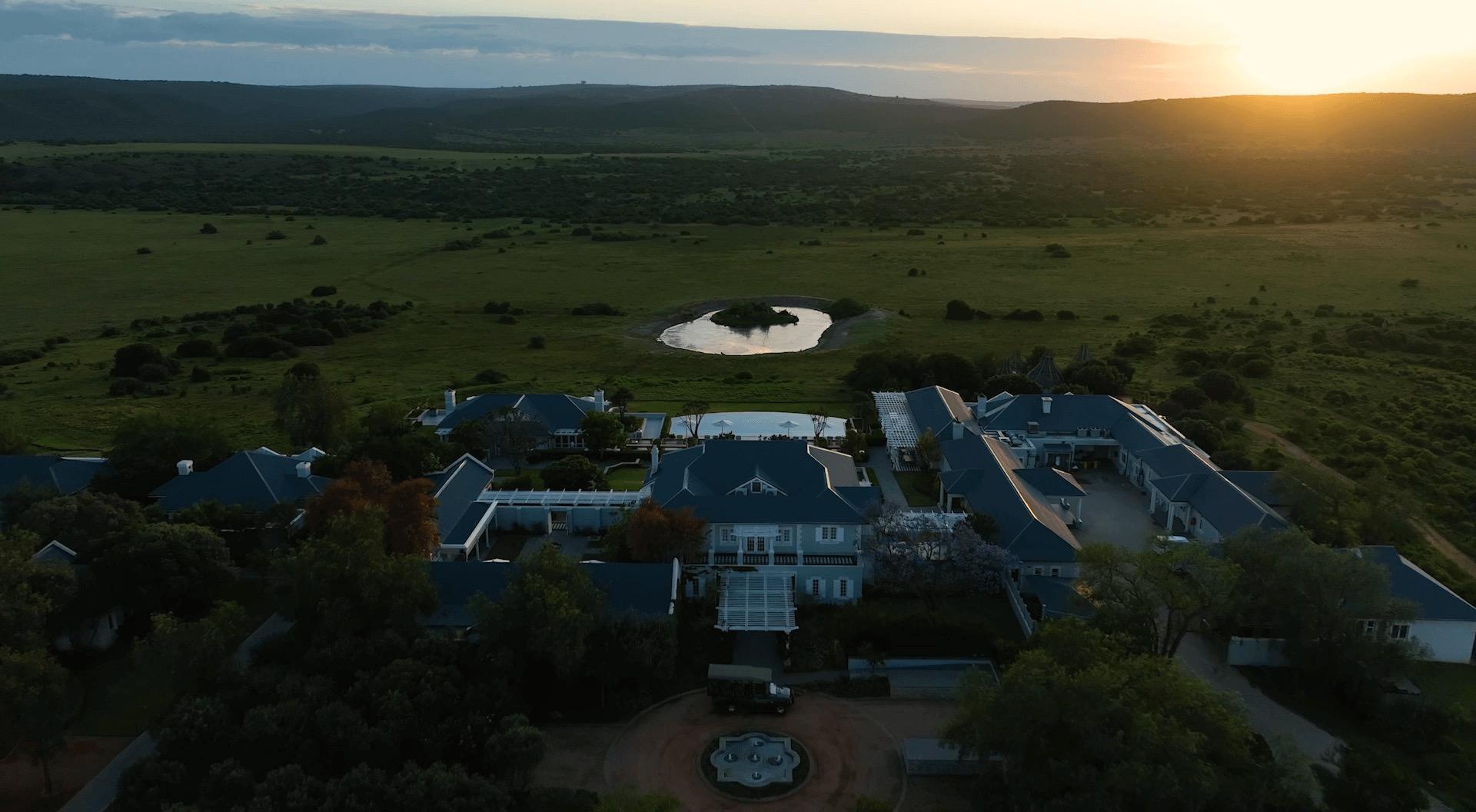 An aerial view at dusk of Long Lee Manor's white colonial buildings, a pool and a round dam, set among grassland and hills.