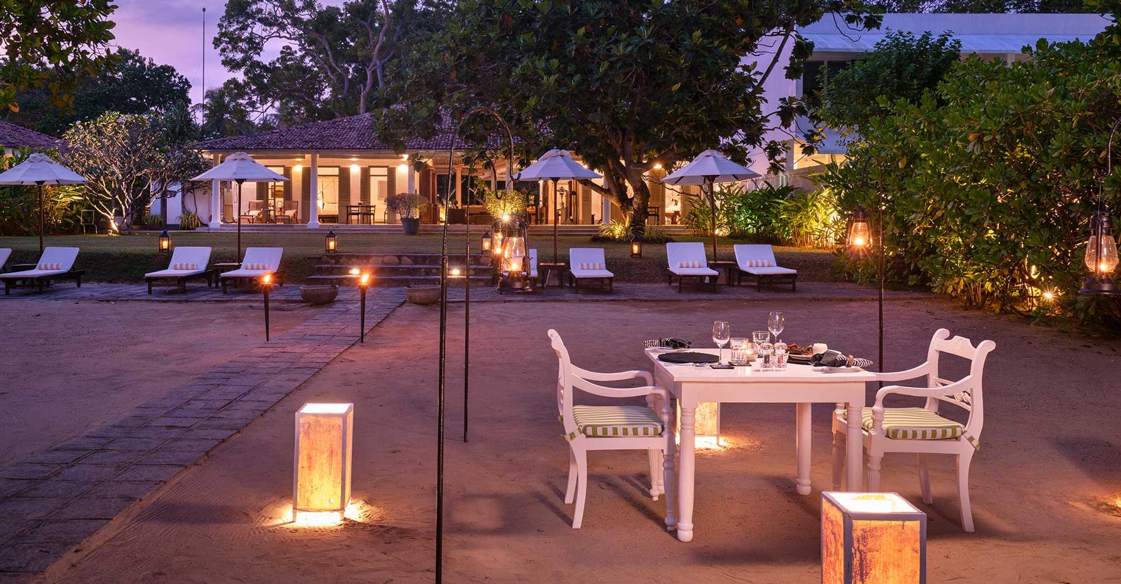 A table set for two on sand at dusk, with wine glasses, striped chairs and lanterns on the ground, and a white pavilion behind.