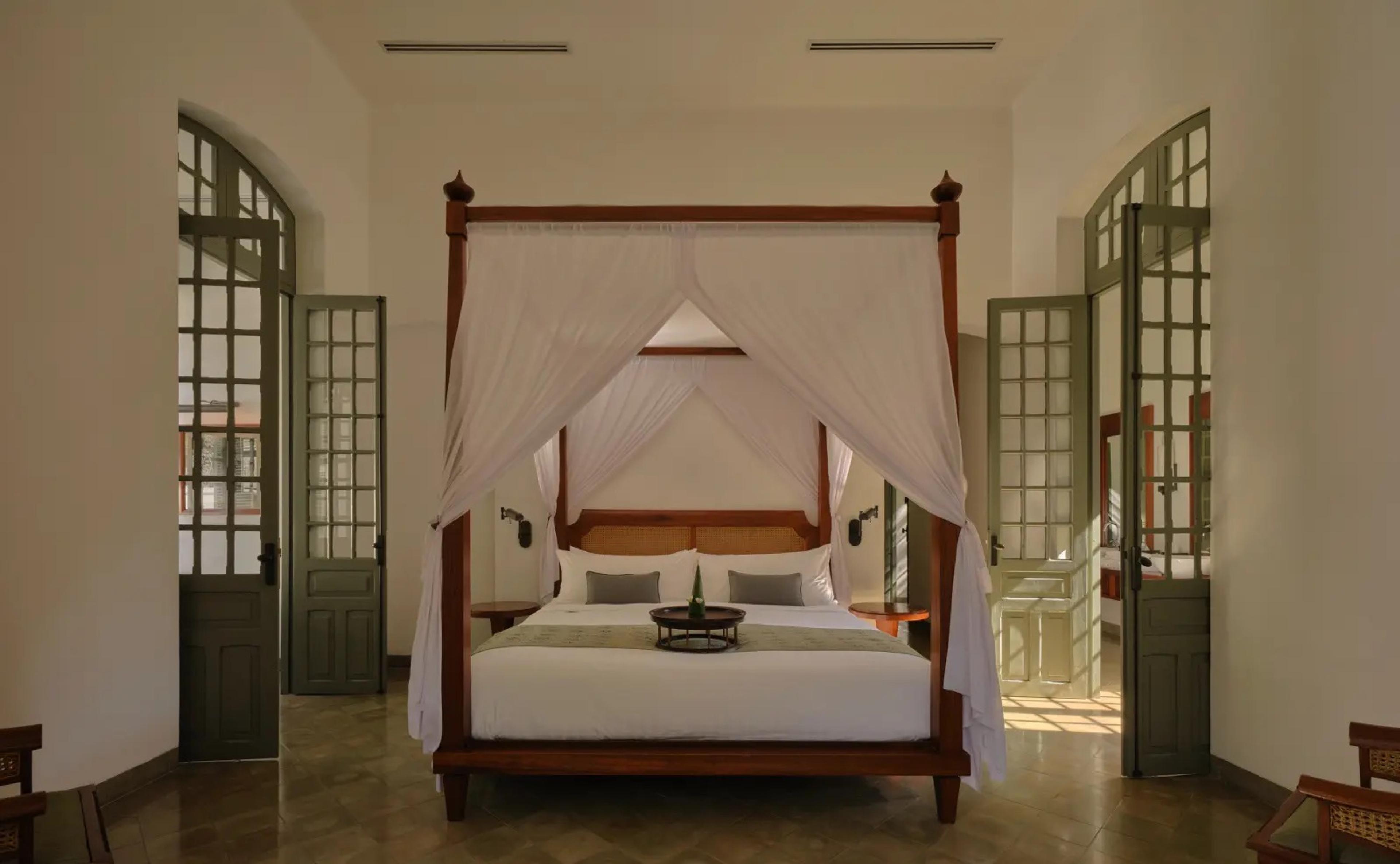 A four-poster bed hung with sheer white curtains in a high-ceilinged room, a drinks tray on the bedspread and doors open to both sides.