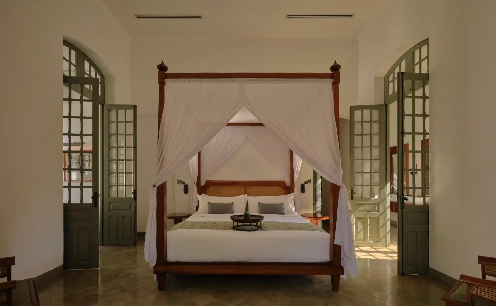 A four-poster bed hung with sheer white curtains in a high-ceilinged room, a drinks tray on the bedspread and doors open to both sides.